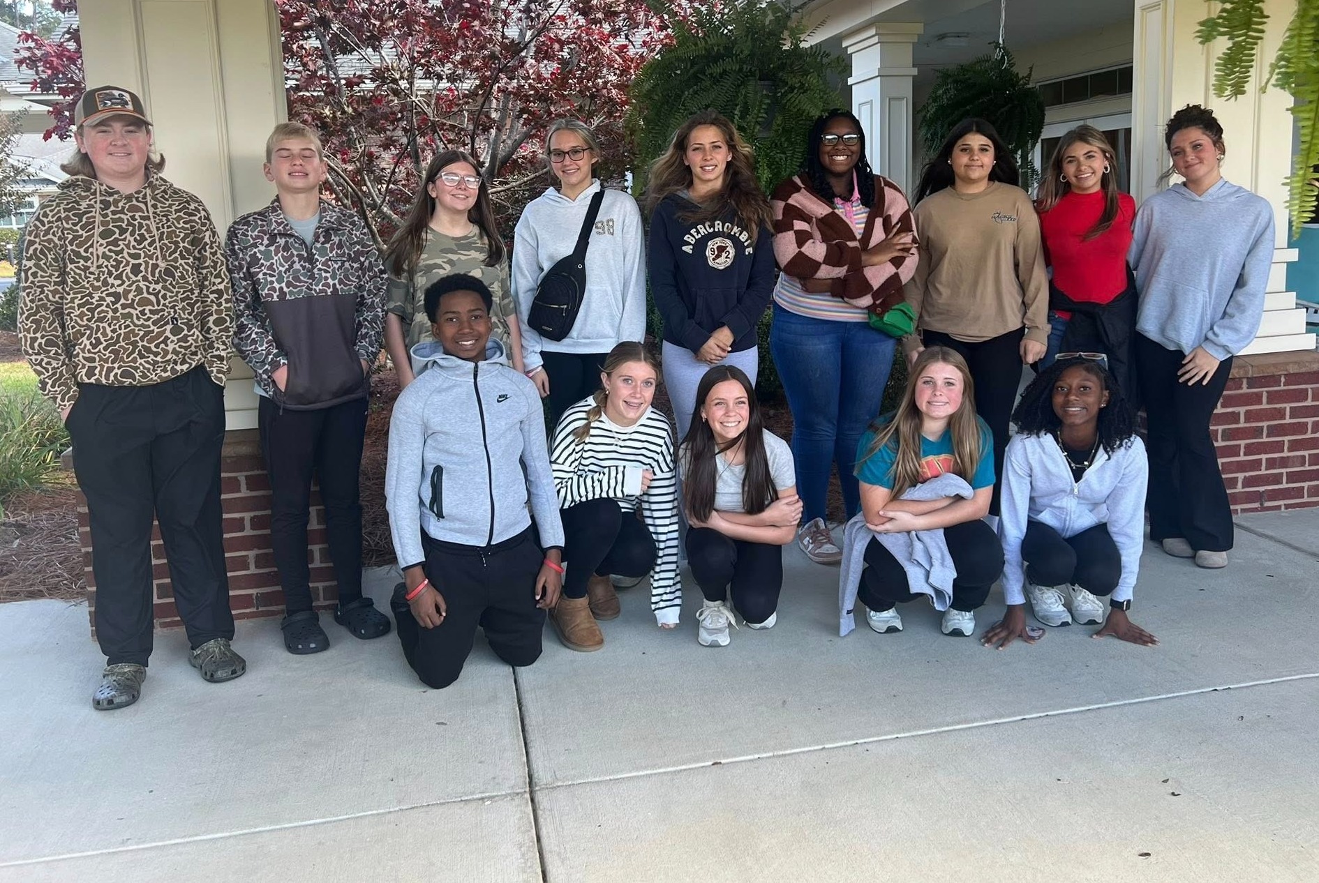 Group of students pose together outside a school building entrance, standing and kneeling under a covered walkway.