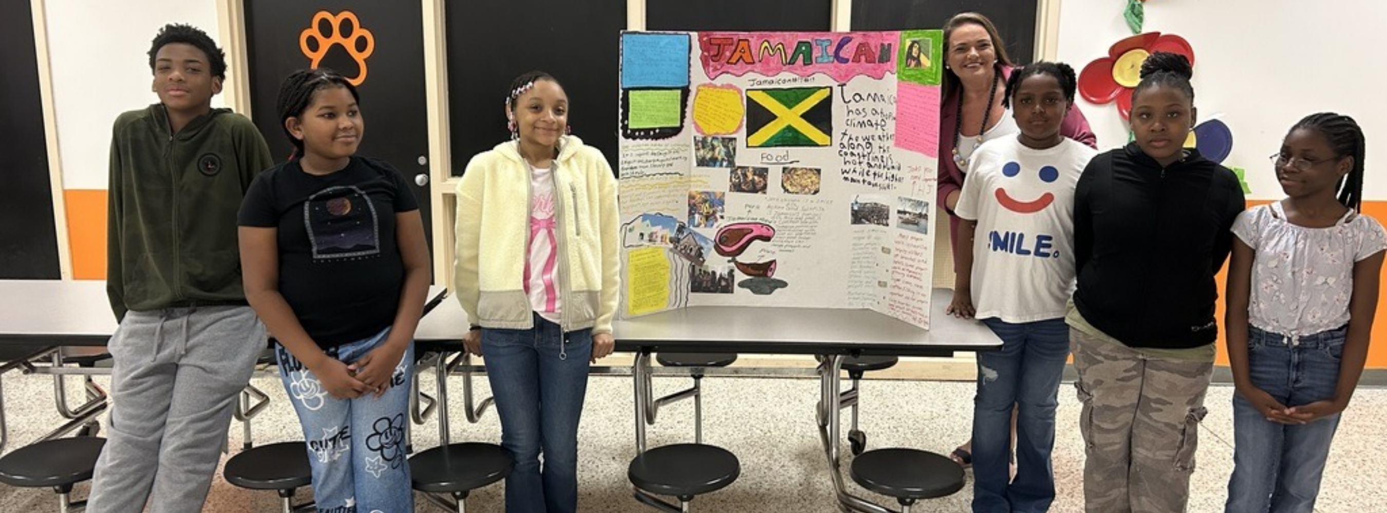 A group of seven students and one female teacher posing in a school hallway with a colorful tri-fold presentation board titled "JAMAICAN." The board features the Jamaican flag, photos of food, and handwritten facts about the country's climate and culture. The students are dressed in casual school attire, and a bright red flower decoration is visible on the wall to the right.