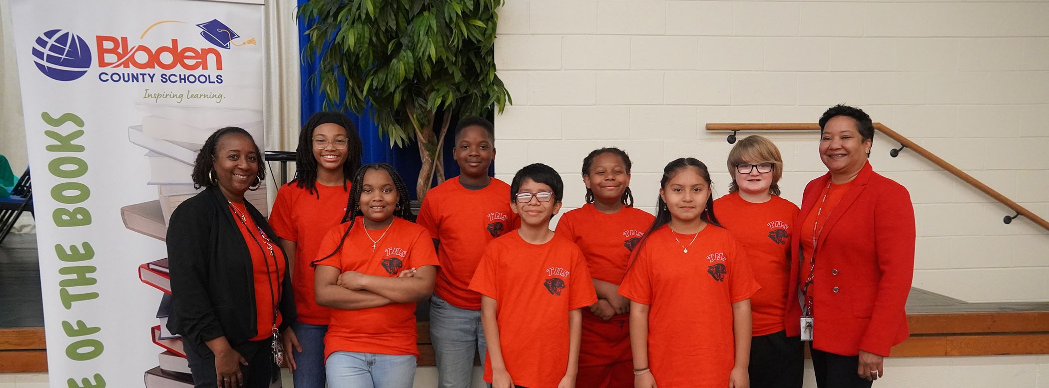 Group of students and two staff members in matching red Tar Heel School t shirts, standing in front of a stage. There is a banner with the BCS logo at the top, that says Battle of the Books.