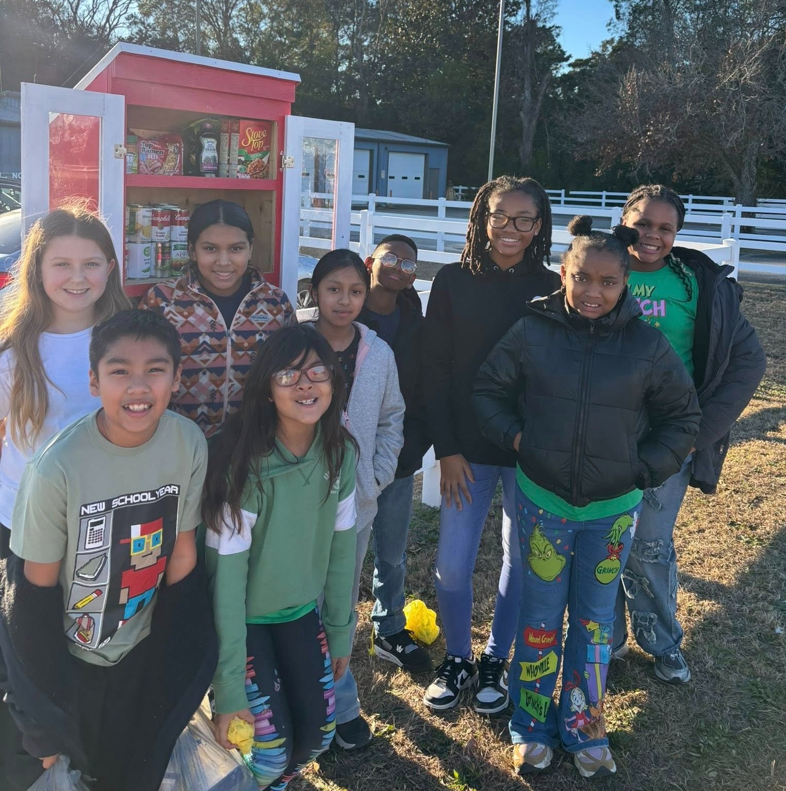 Nine diverse children, ages ranging from elementary to middle school, smile and pose outdoors around a small red-and-white two-door food pantry stocked with nonperishable items (visible labels include Stove Top, Campbell’s, Green Giant). Behind them are trees, a white fence, and a building with garage doors. In the front left, a boy’s shirt says “NEW SCHOOL YEAR” with school-themed icons; a girl in green wears Grinch-themed jeans and a shirt partially reading “MY GRINCH.”