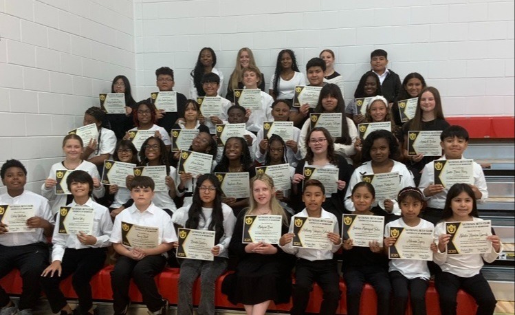 Group photo of about 45–50 middle school students arranged in four tiered rows inside a school gymnasium, each holding a certificate with a gold seal identifying them as members of the National Beta Club, Junior Division (2023–2024). Most students wear white or black tops with dark bottoms. The background shows white brick walls with red and gray panels and a wooden basketball court floor with a red painted area.