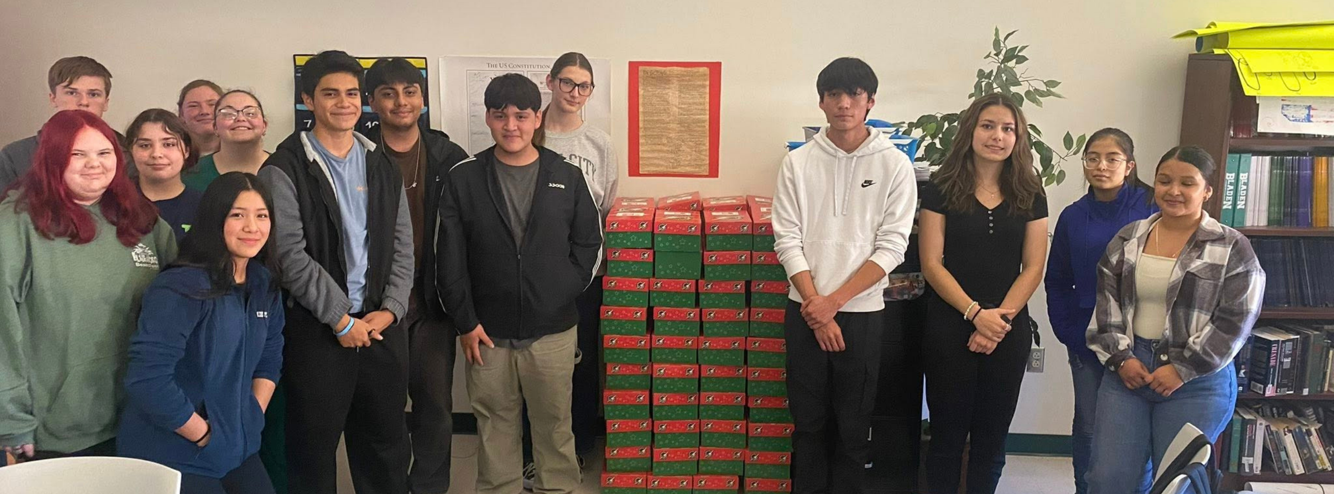 A group of thirteen high school students stands in a classroom, posing behind a large, neat stack of red and green gift boxes. The classroom walls are decorated with educational posters, including one of the U.S. Constitution, and a bookshelf filled with binders is visible on the right. The students are dressed casually in hoodies and t-shirts, appearing proud of their collection effort.
