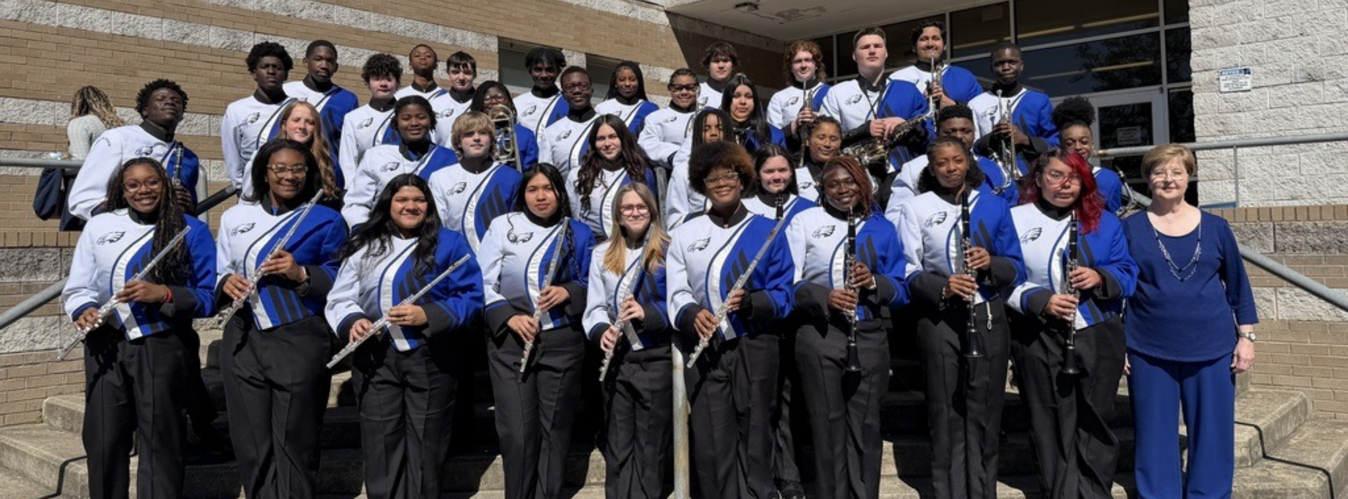 A wide group portrait of the East Bladen High School marching band posed outdoors on concrete steps in front of a brick building. The students are wearing professional blue, white, and black uniforms featuring an eagle logo on the chest. Many students in the front rows are holding instruments, including flutes and clarinets, while students in the back rows hold brass instruments and saxophones. An adult instructor in a blue blouse stands on the far right.