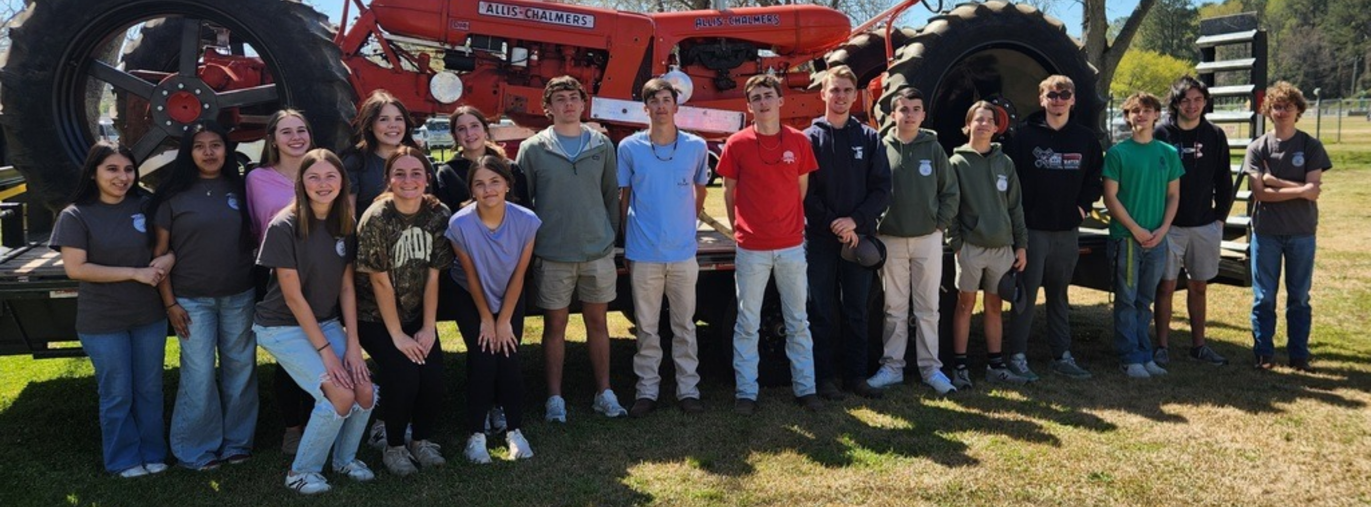 A wide shot of a large group of students posing outdoors in front of a vintage orange Allis-Chalmers tractor parked on a flatbed trailer. The students are dressed casually, and the scene suggests a project completion or a "Tractor Day" event on campus.
