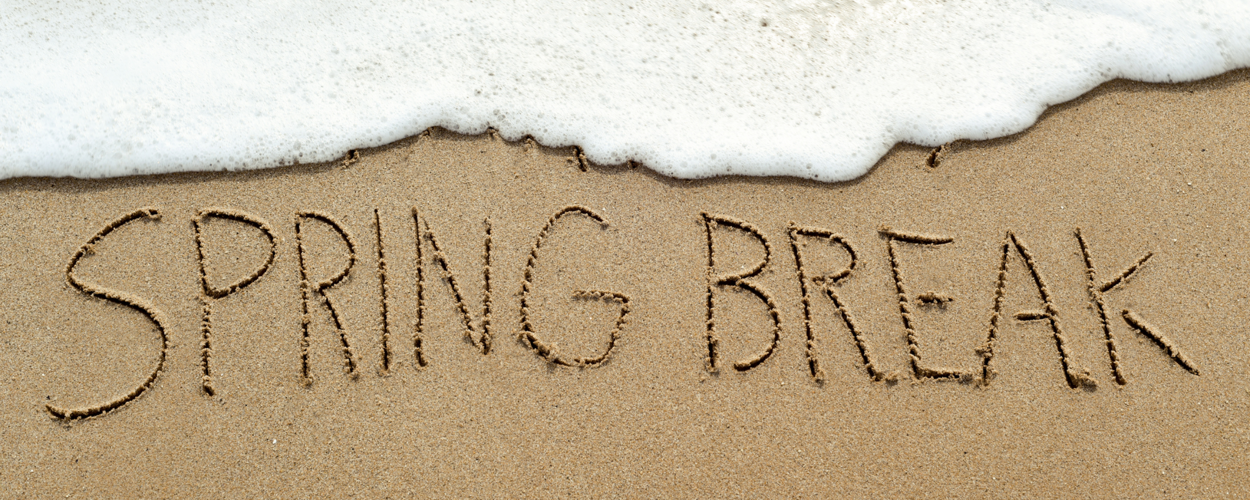 A wave crashes over the beach, in the sand it says "Spring Break"