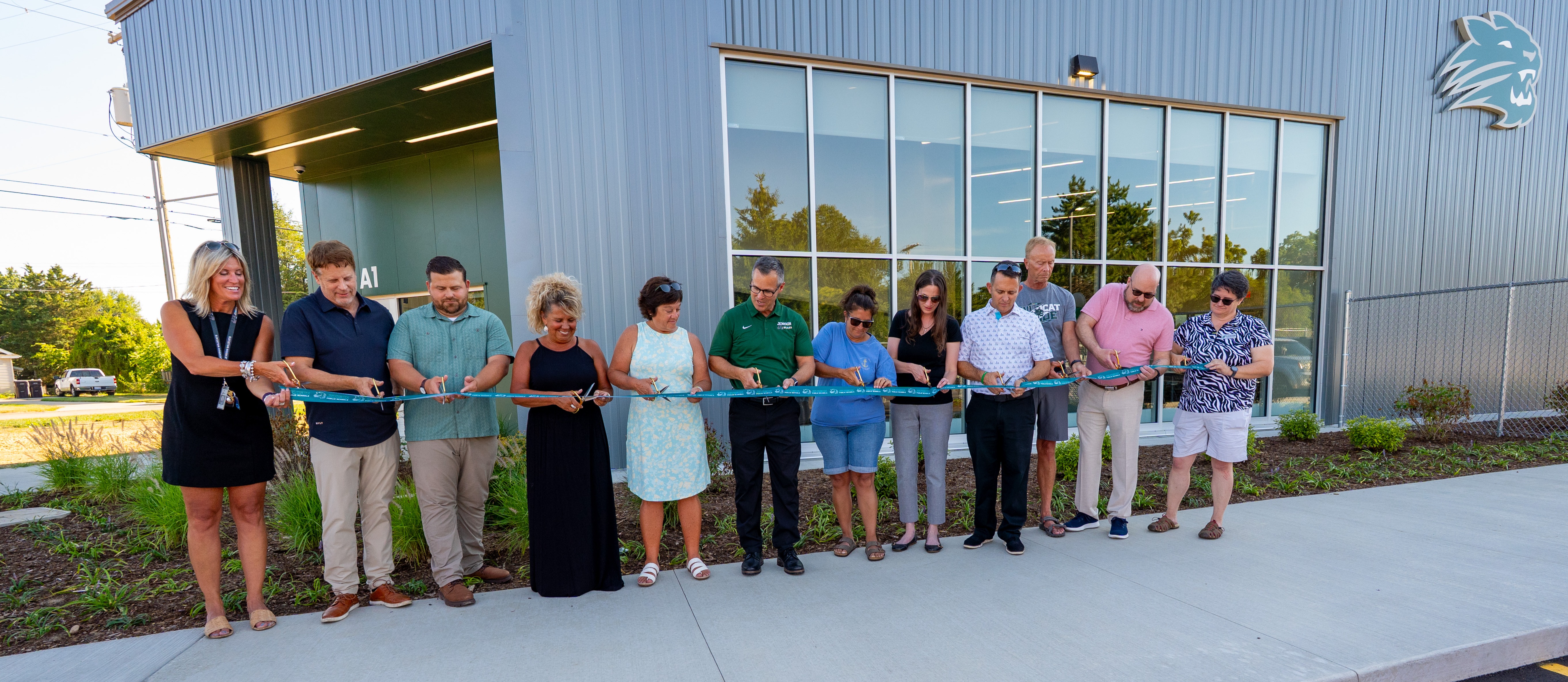 People holding a ribbon to be cut stand in line in front of a building.