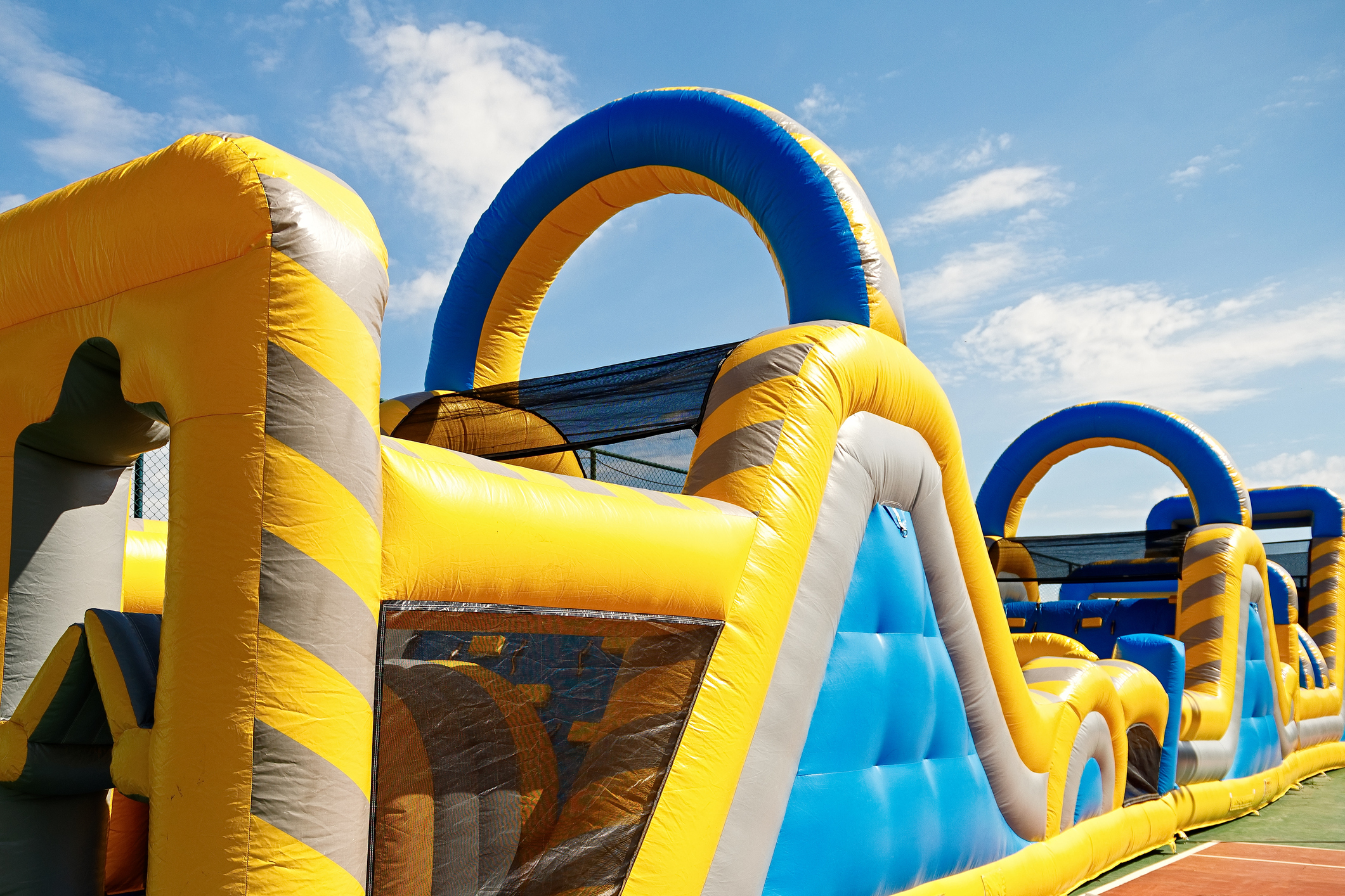 Large inflatable obstacle course with yellow and blue sections. Features a net, slide, and open spaces. Set against a bright blue sky with some clouds.