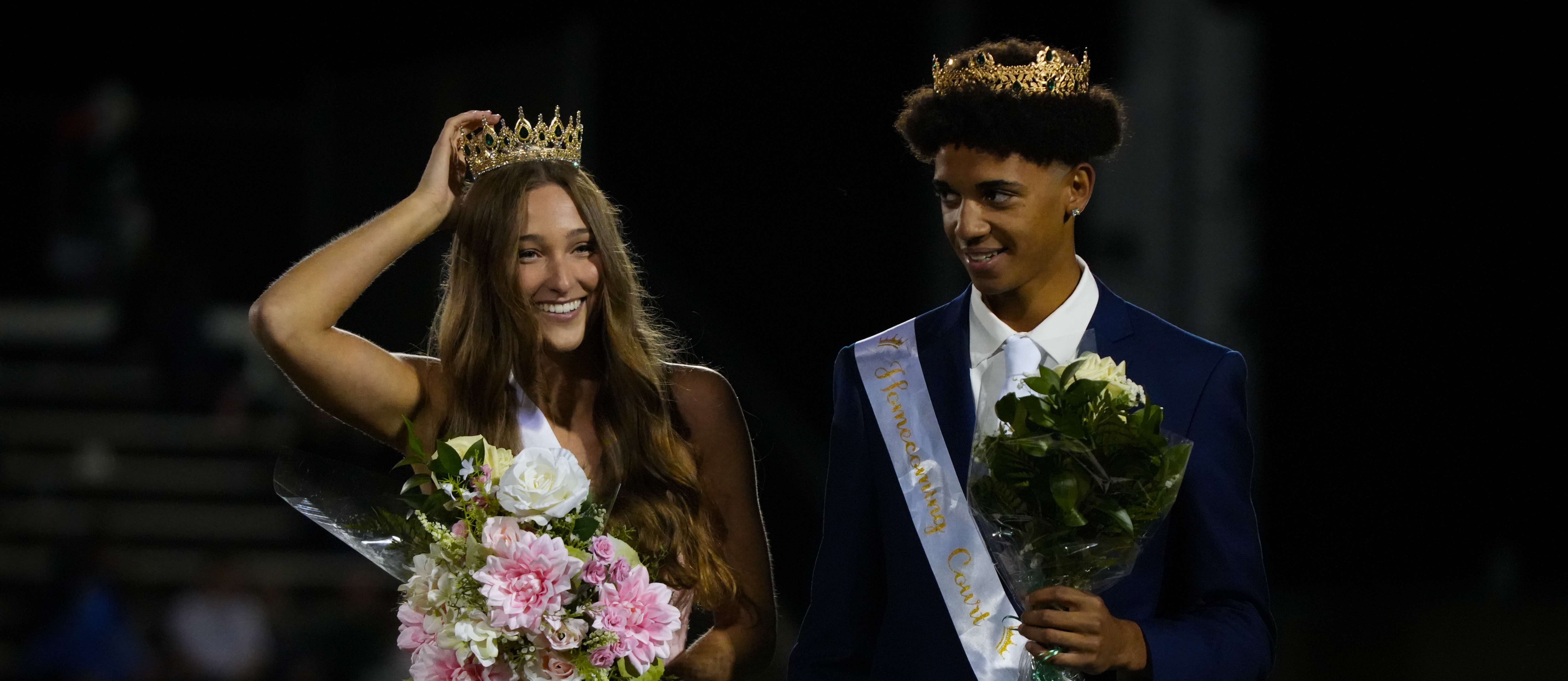 A smiling crowned woman in pink and a suited man in blue hold flowers, standing on a stage.