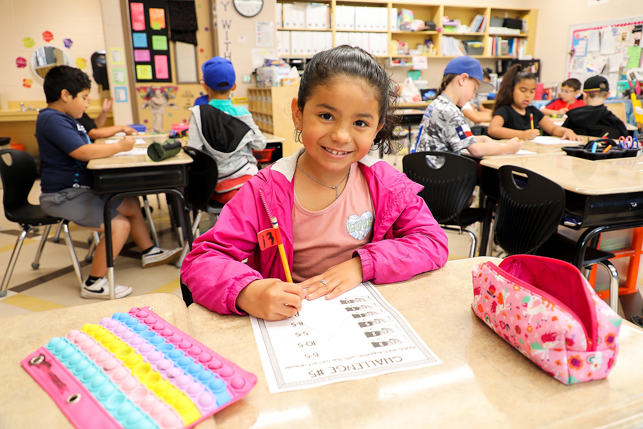 Elementary student in a pink jacket smiles while completing a classroom worksheet at her desk. Other students are seated and working independently in the background of the brightly lit classroom.