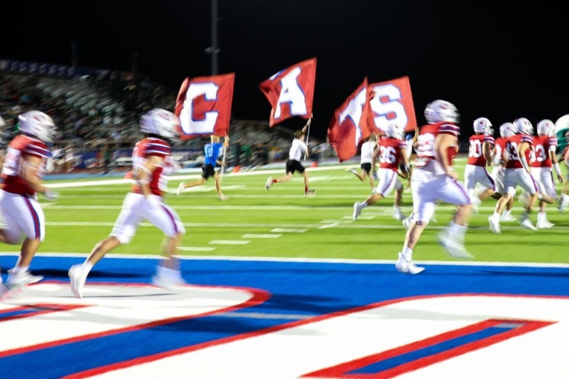Gregory-Portland Wildcats football players run onto the field during a nighttime game carrying team flags as they take the field under stadium lights.