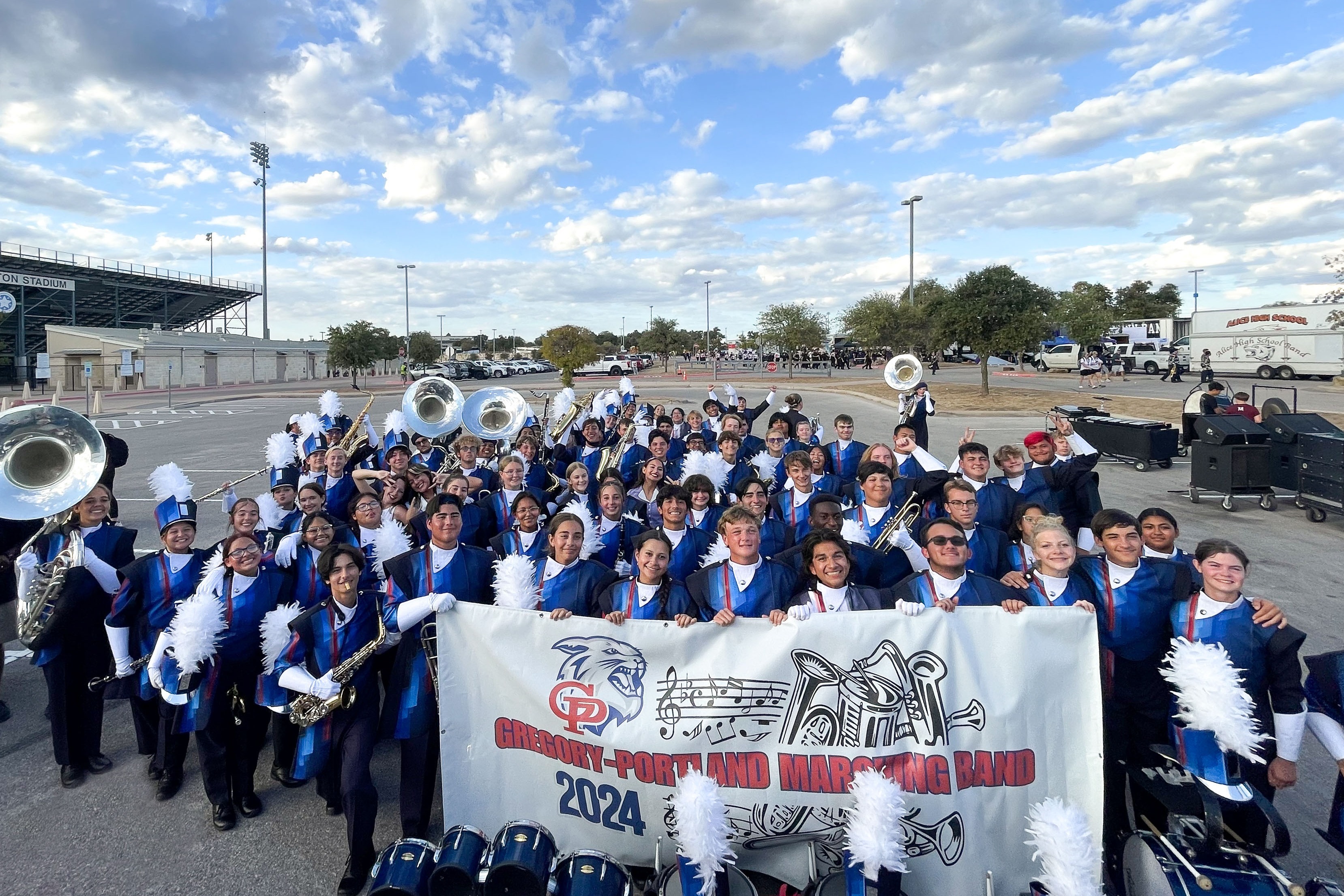 Members of the Gregory-Portland High School Marching Band pose for a group photo in uniform while holding a band banner outside a stadium before a performance.