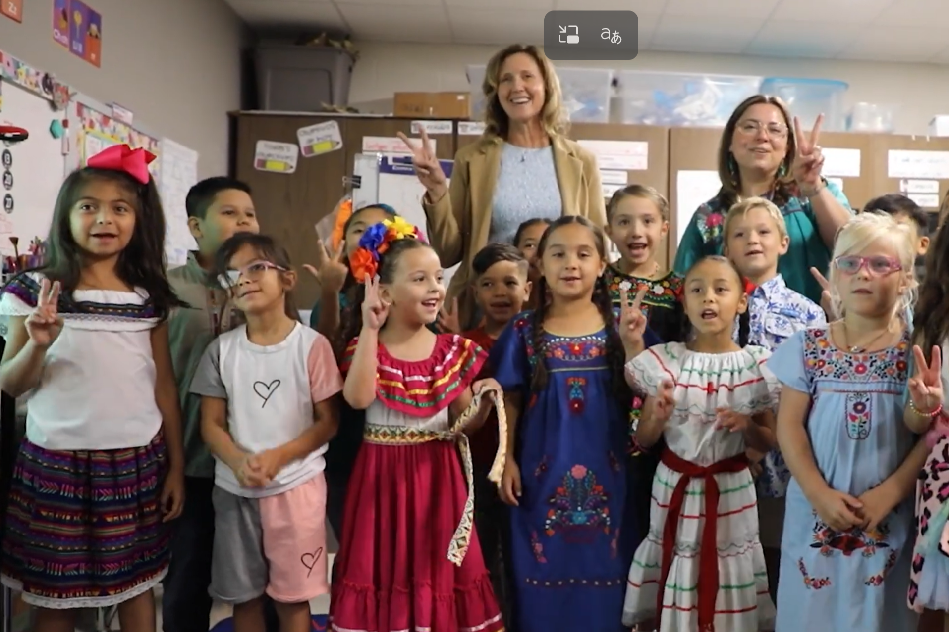 Elementary students and two teachers pose together in a classroom, many wearing colorful cultural attire and holding up peace signs.