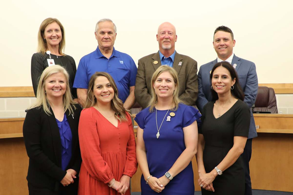 Group portrait of eight school district leaders and board members standing in a boardroom. Four individuals stand in the front row and four in the back row, smiling toward the camera. The group is positioned in front of a board meeting dais, with members dressed in professional attire including business suits, dresses, and collared shirts. The photo appears to have been taken during a formal board or district leadership meeting.