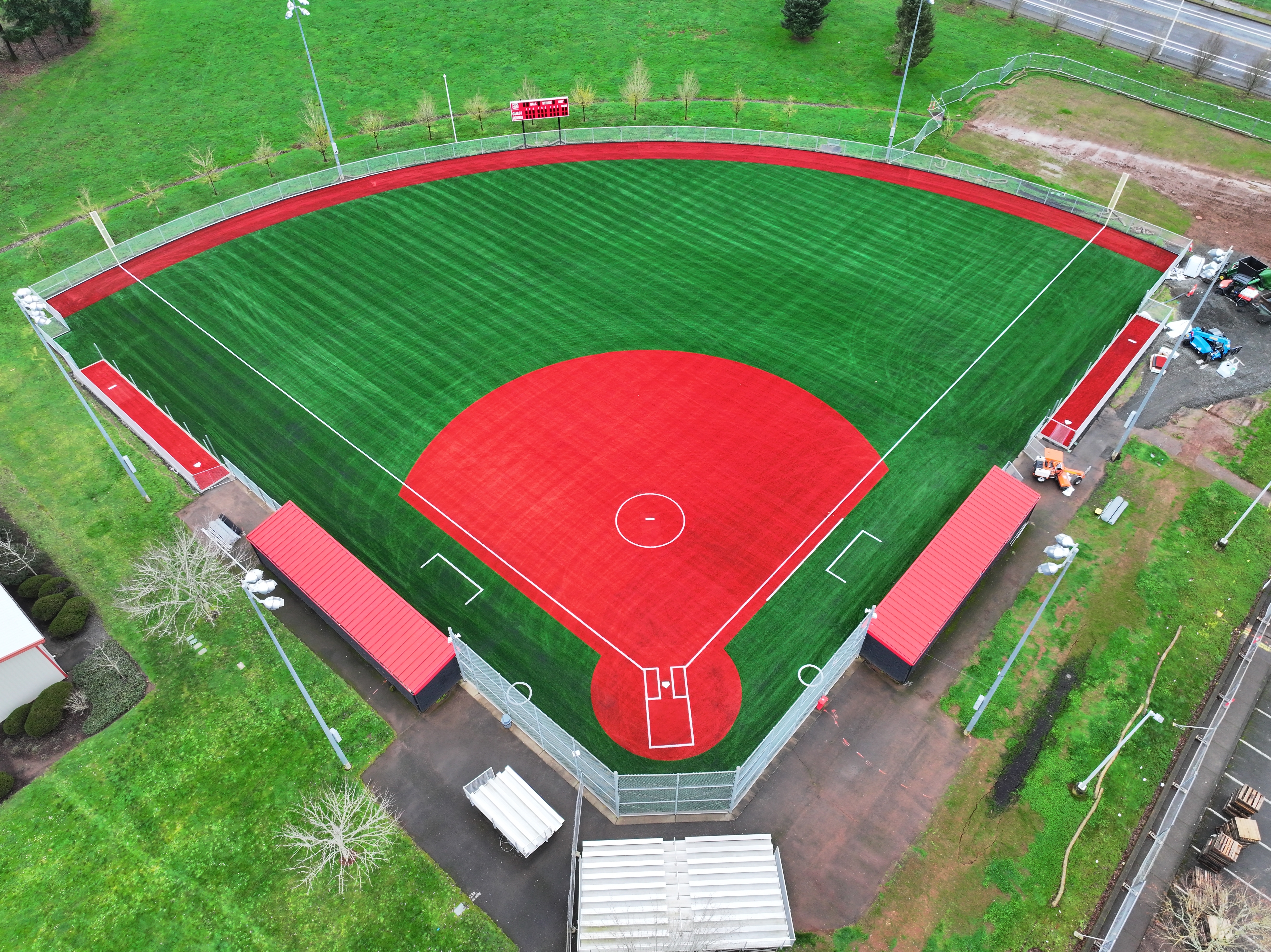 Aerial drone view of a newly installed turf softball field at Oregon City High School. The image shows bright green artificial turf with red infield and warning track areas, clearly marked baselines and pitcher’s circle, dugouts on both sides, fencing, bleachers behind home plate, and surrounding grassy areas