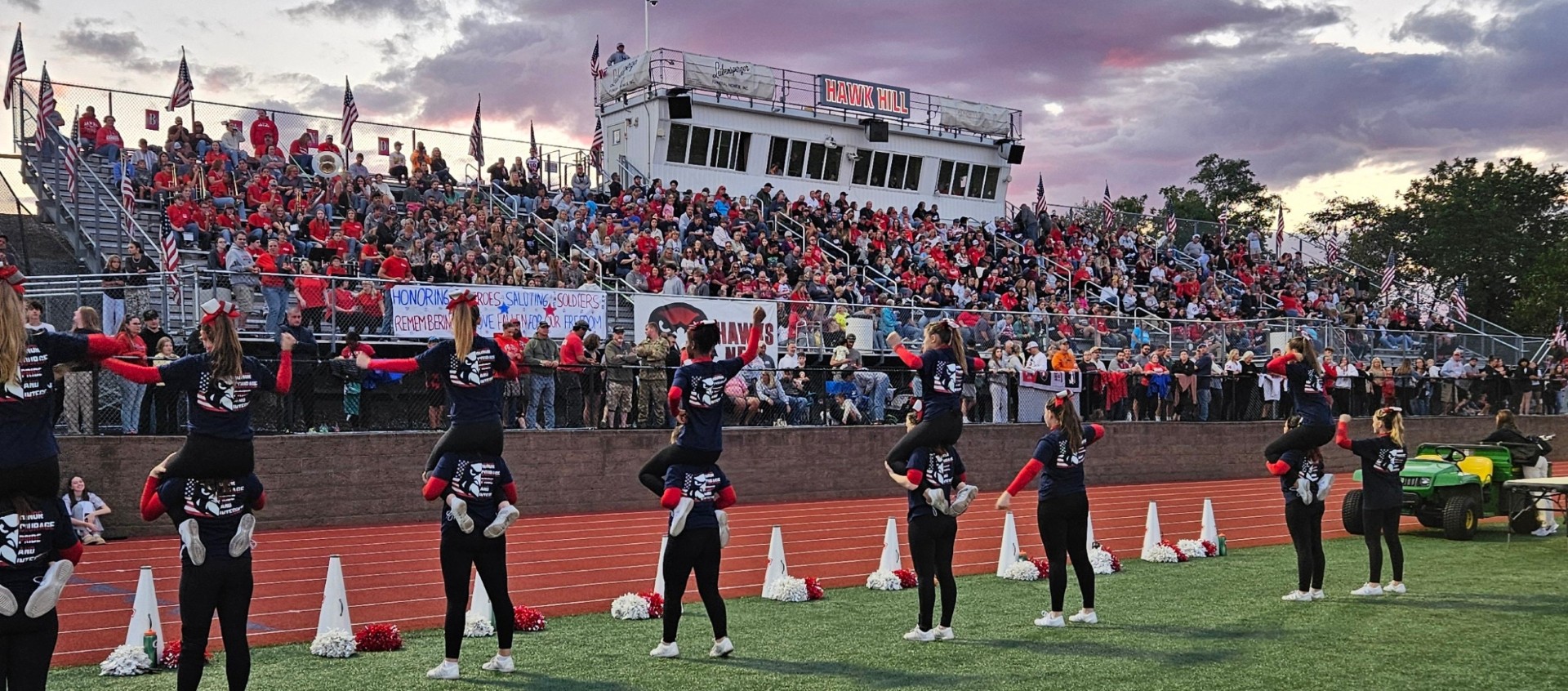 The Hawk Hill stadium bleachers packed with onlookers with American flags around the bleachers and the Cheer team performing an exciting cheer routine.