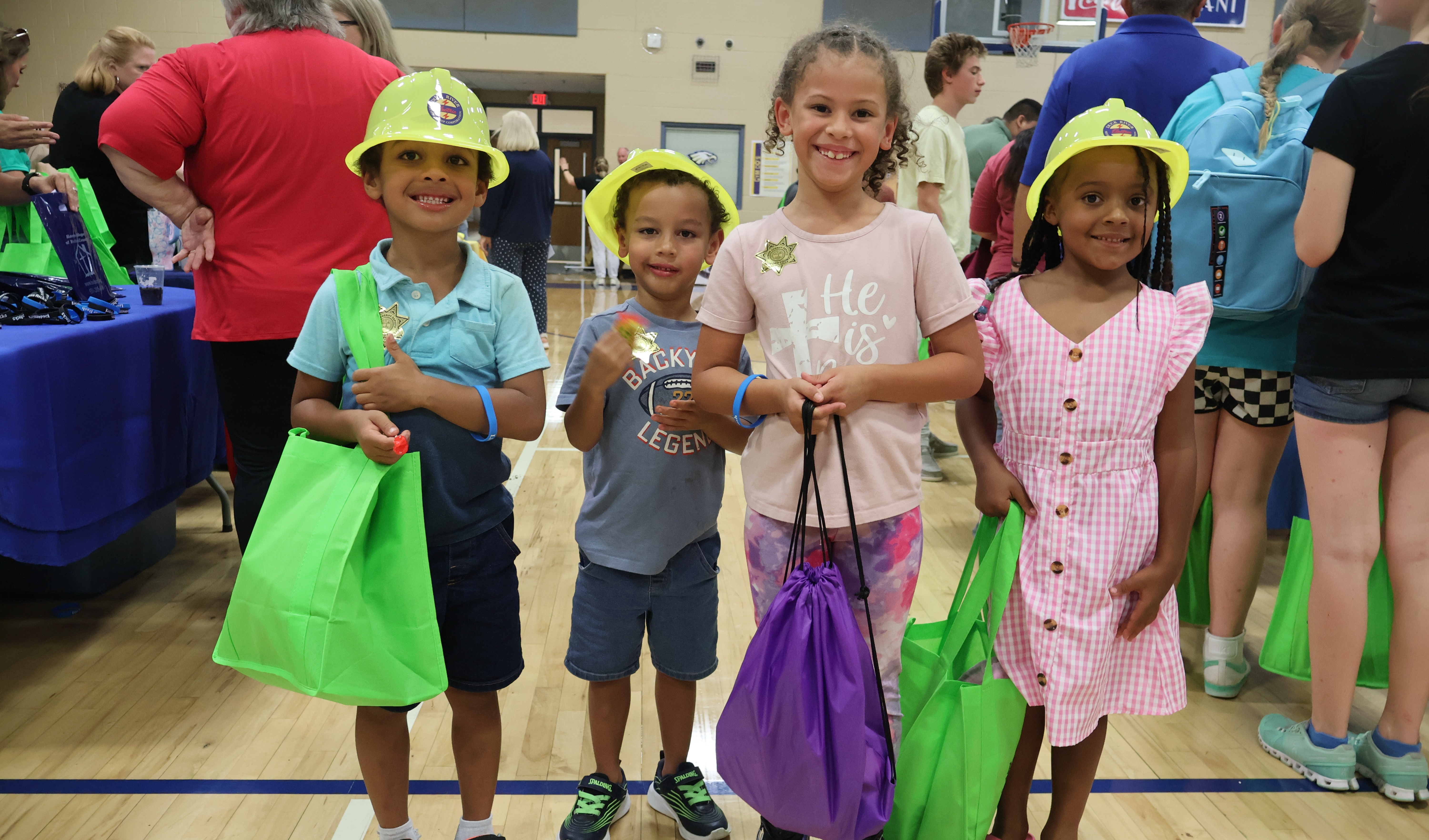There are four children posing for a picture inside a busy gym each holding up things that they were given from the event. 