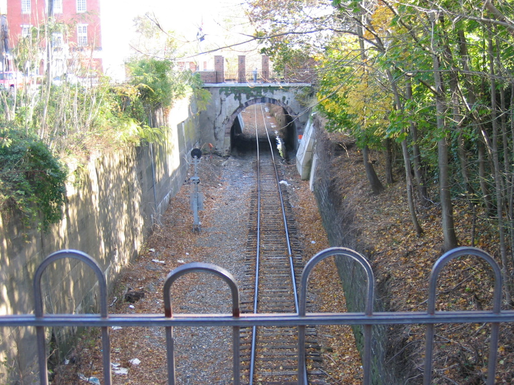 A view of train tracks leading into a tunnel, seen from behind a metal fence, with trees and buildings in the background.