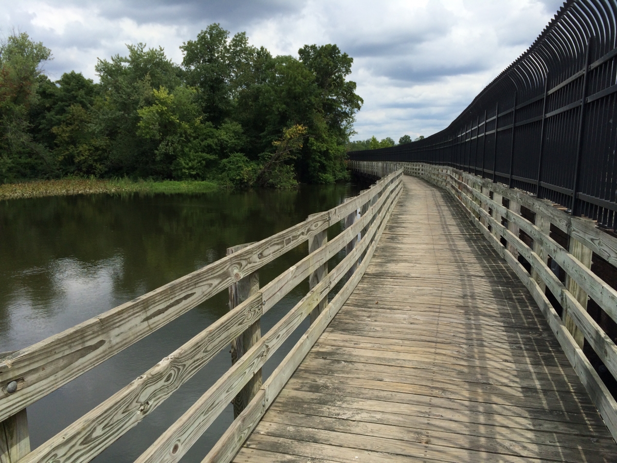 A wooden walkway with railings runs alongside a river, bordered by trees and a black fence on the right, under a cloudy sky.