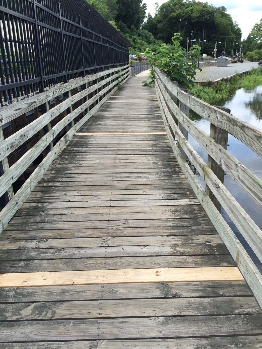 A wooden pedestrian bridge with railings on both sides, crossing over a body of water with trees and a road in the background.