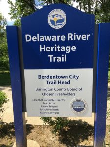 A blue sign indicating the Delaware River Heritage Trail at Bordentown City Trail Head, with trees in the background.