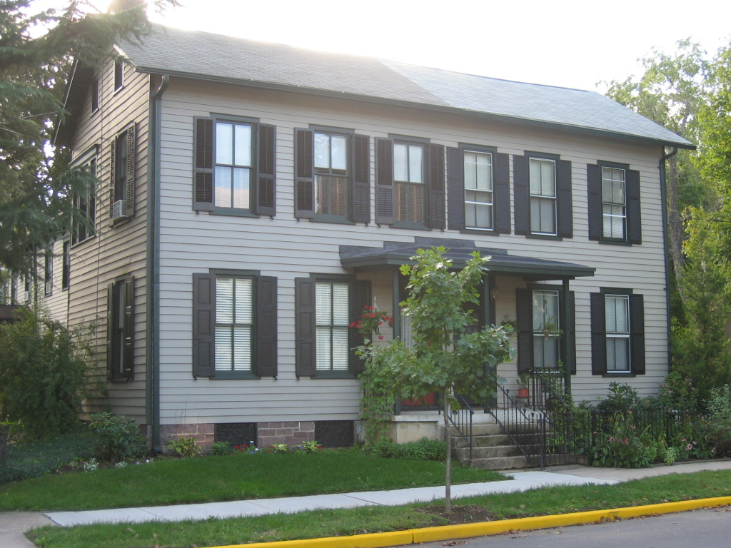 A two-story house with beige siding, black shutters, and a small front porch with steps, surrounded by a lawn and trees.