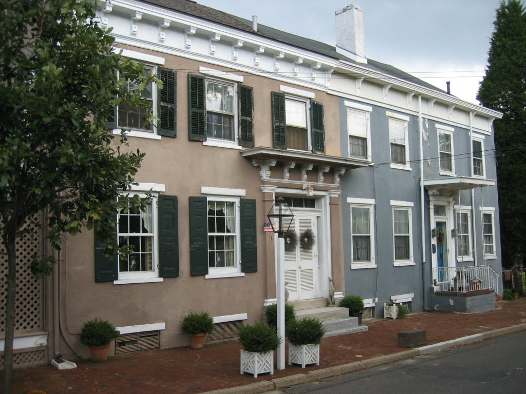Two adjacent row houses with different colors, green shutters, and small front porches, on a brick sidewalk.