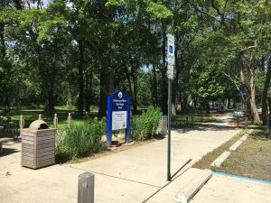 A park entrance with a blue sign, a trash bin, and a parking meter, surrounded by trees and greenery.