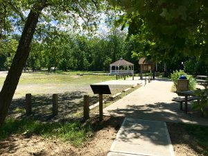 A park scene with a paved walkway, benches, trees, and a small pavilion in the background.