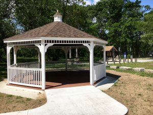 A white wooden gazebo with a shingled roof in a park, surrounded by trees and a paved walkway.