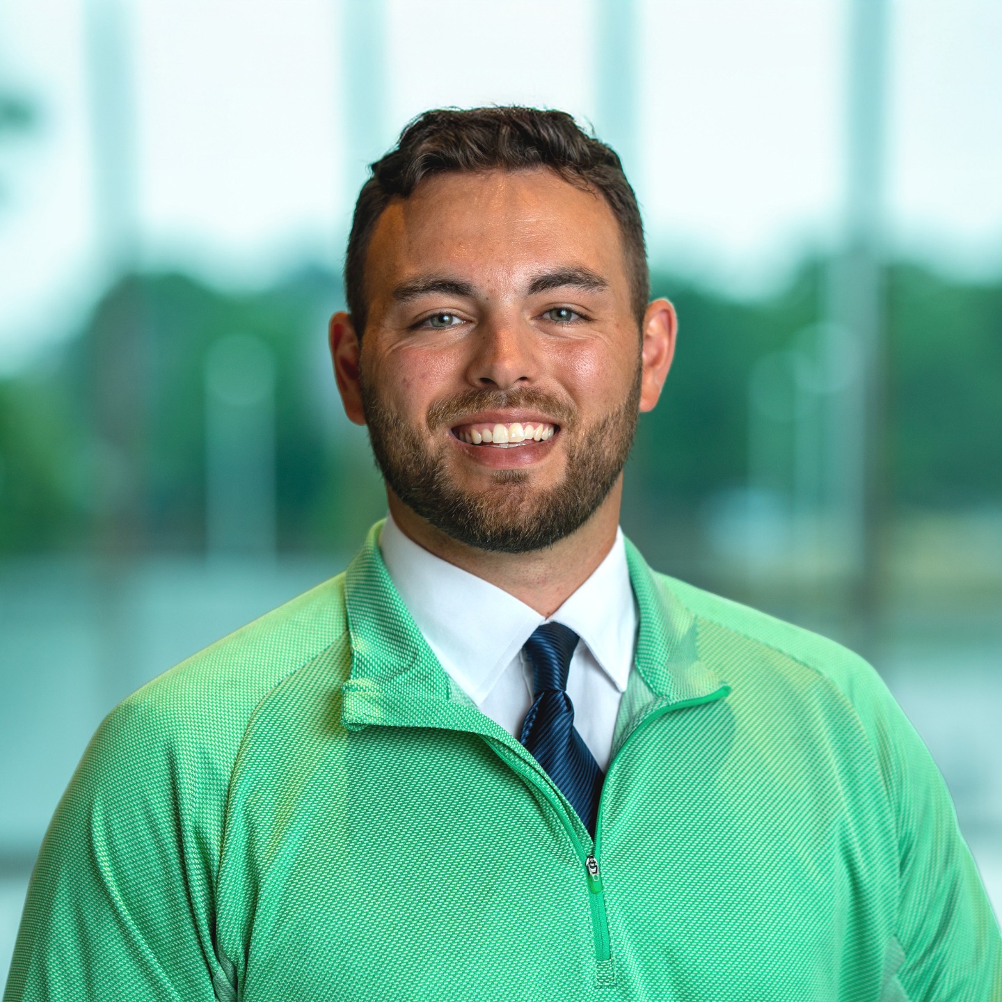A man with a beard wearing a green shirt and blue tie smiles at the camera with a bright background.