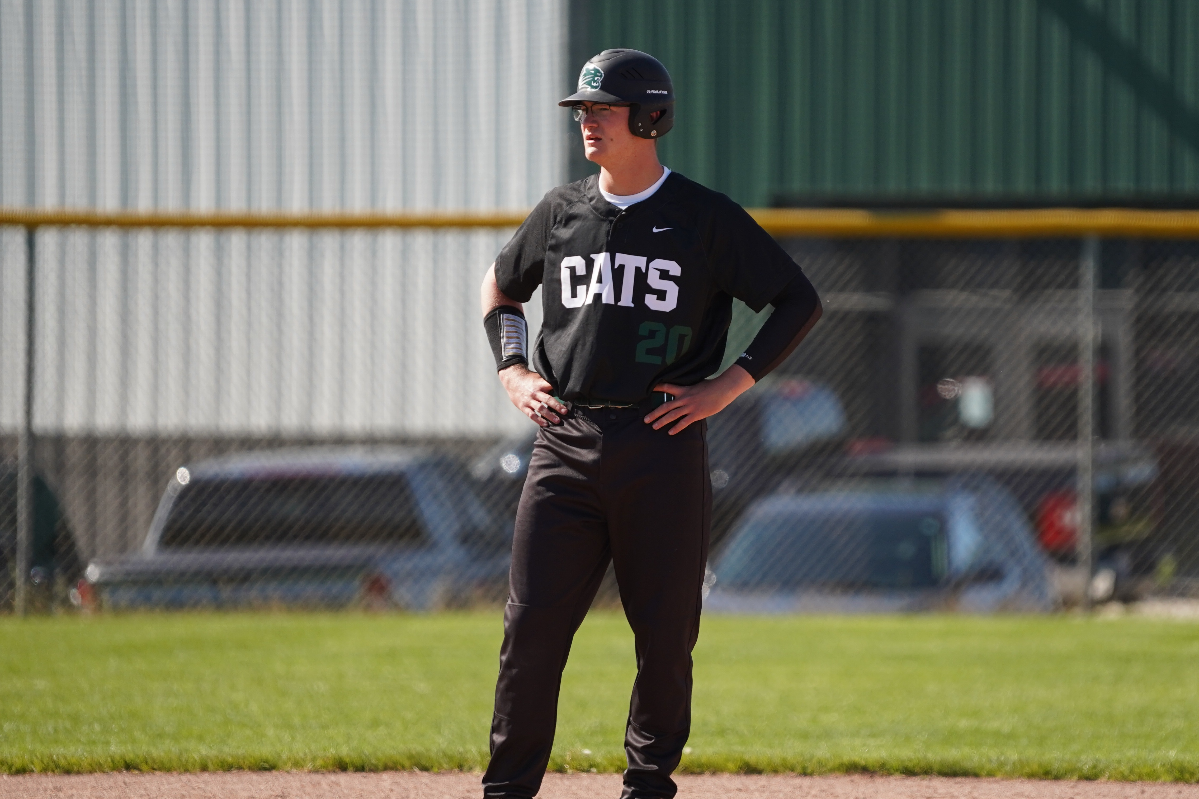 A baseball player stands on base with the field house in the background.