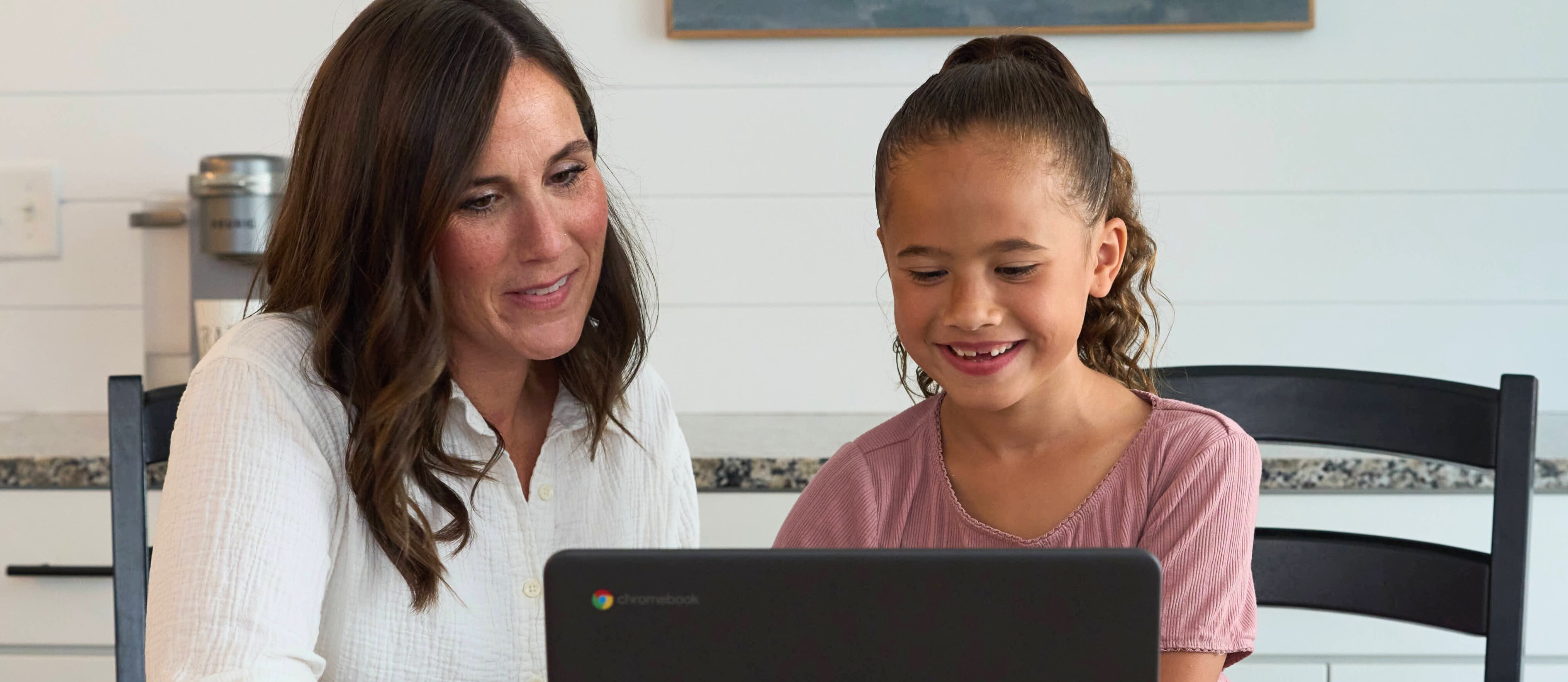 A mother and student sit at a dining room table.  The student works on a computer.