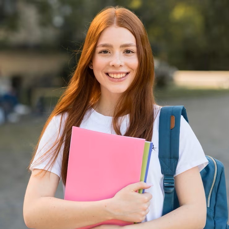 A person holds a pink folder with a smile, wearing a white shirt and carrying a blue backpack.