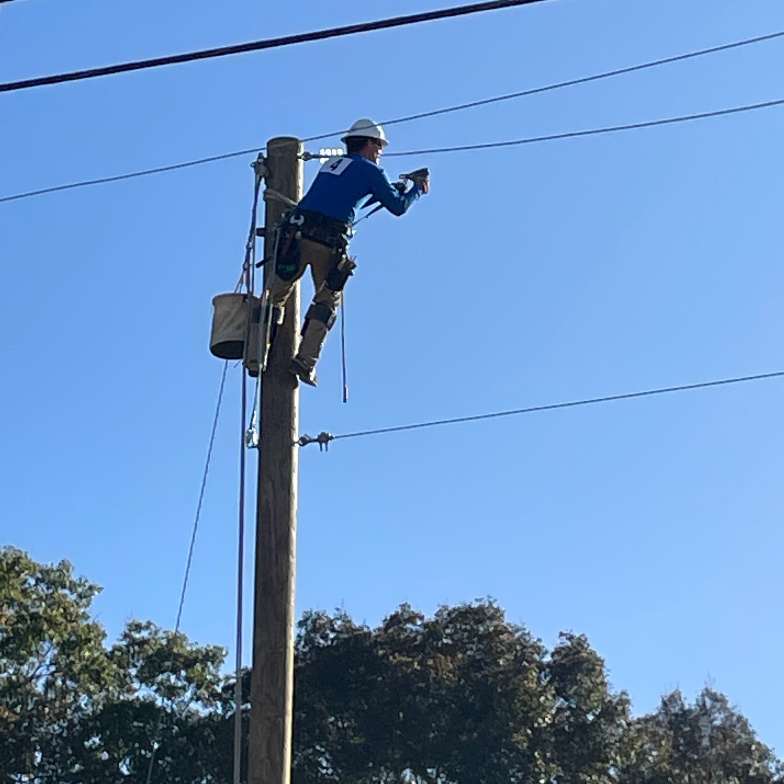 Person in a hard hat and harness climbing a pole with a bucket and tool belt.
