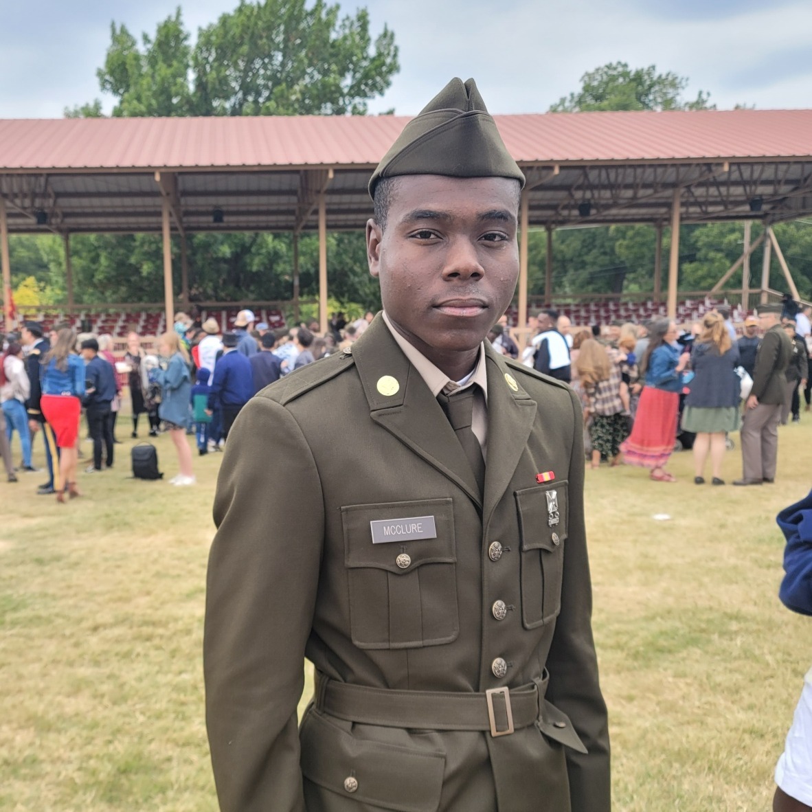 Man in military uniform standing in an outdoor area with a large crowd in the background.