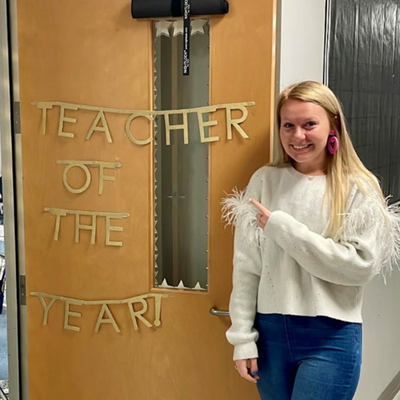 A woman in a white sweater and blue jeans stands in front of a door decorated with a banner.