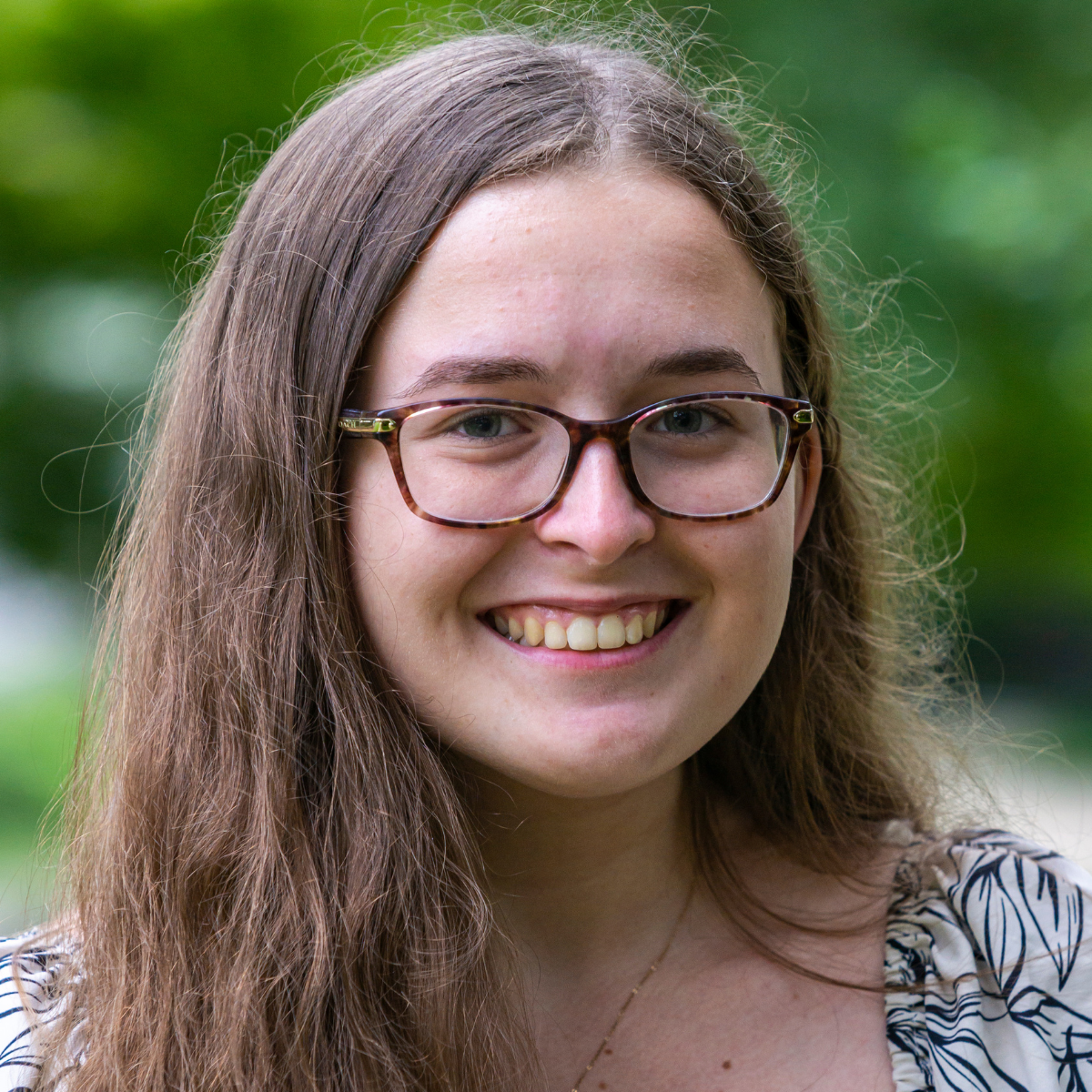 A woman with long, wavy hair, wearing glasses, smiles brightly. She has a necklace and a patterned top.
