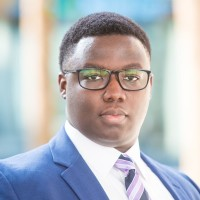 A man in a blue suit and tie with glasses looks at the camera. The background is a blurred office setting.