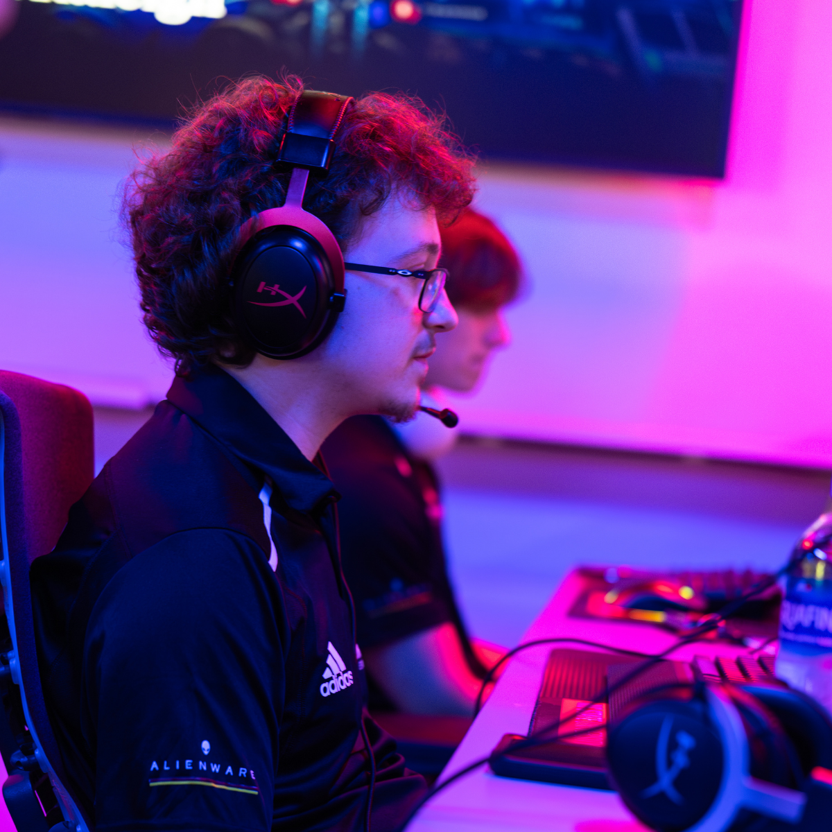Person wearing headphones and glasses at a gaming setup. Another person sits behind them.
