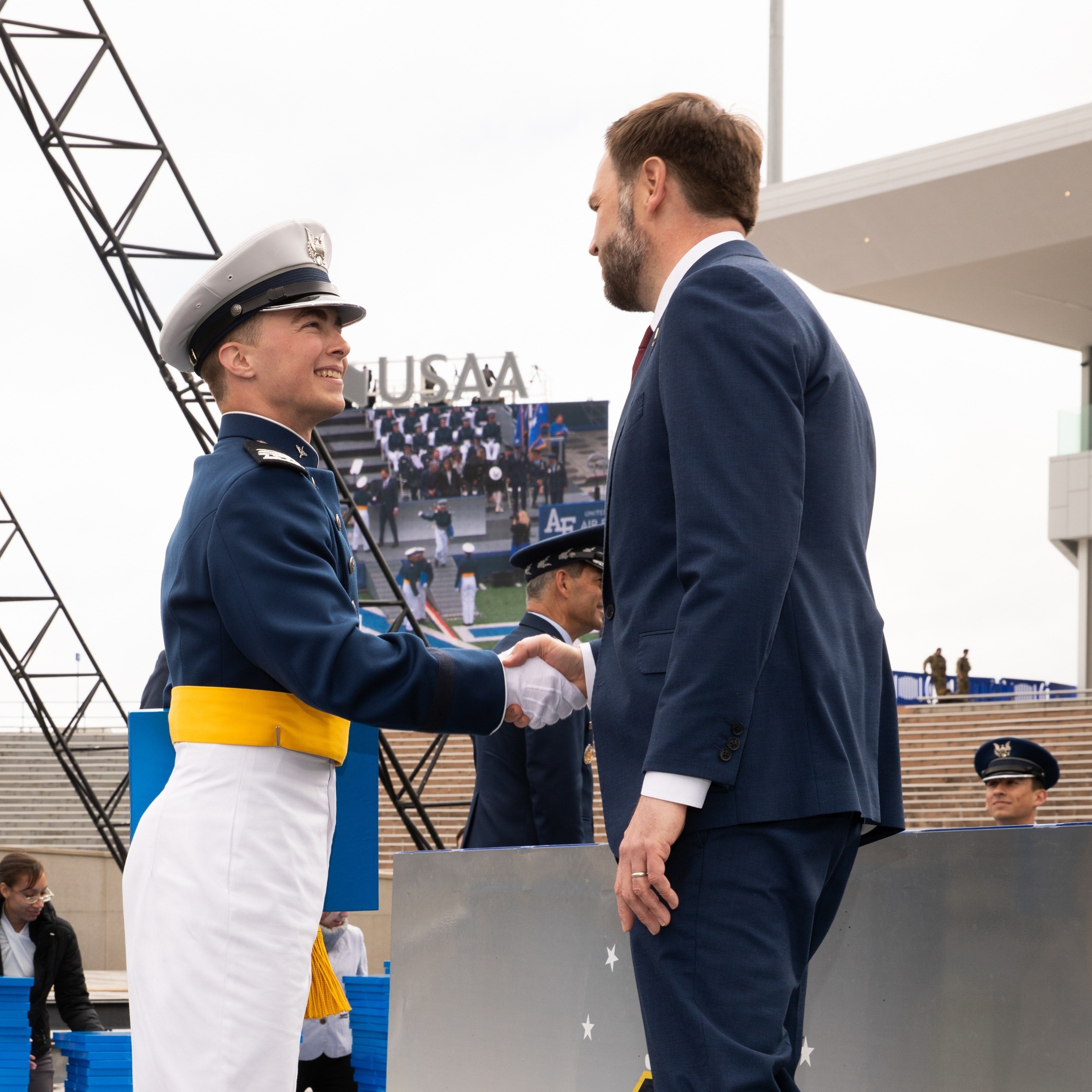 Officer in uniform shaking hands with a man in a suit in front of a large screen.