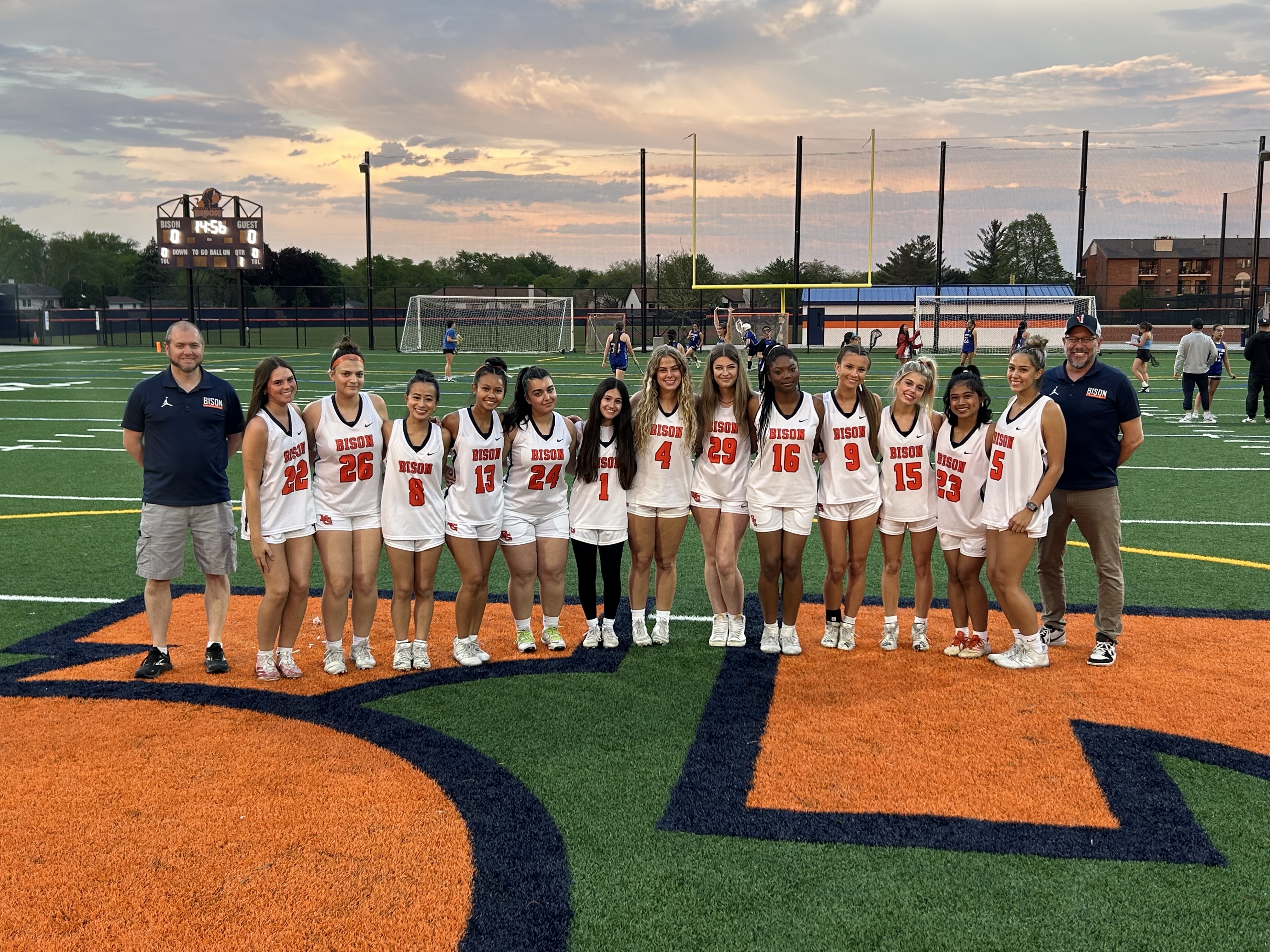 Girls lacrosse team seniors in a line across the center of the turf field with coaches on the end