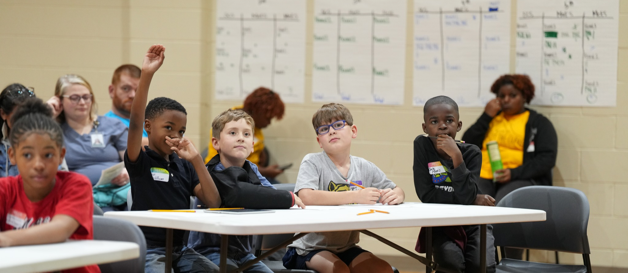This photo has a group of elementary boys participating in the district math quiz bowl. One student is raising his hand to answer a question. 