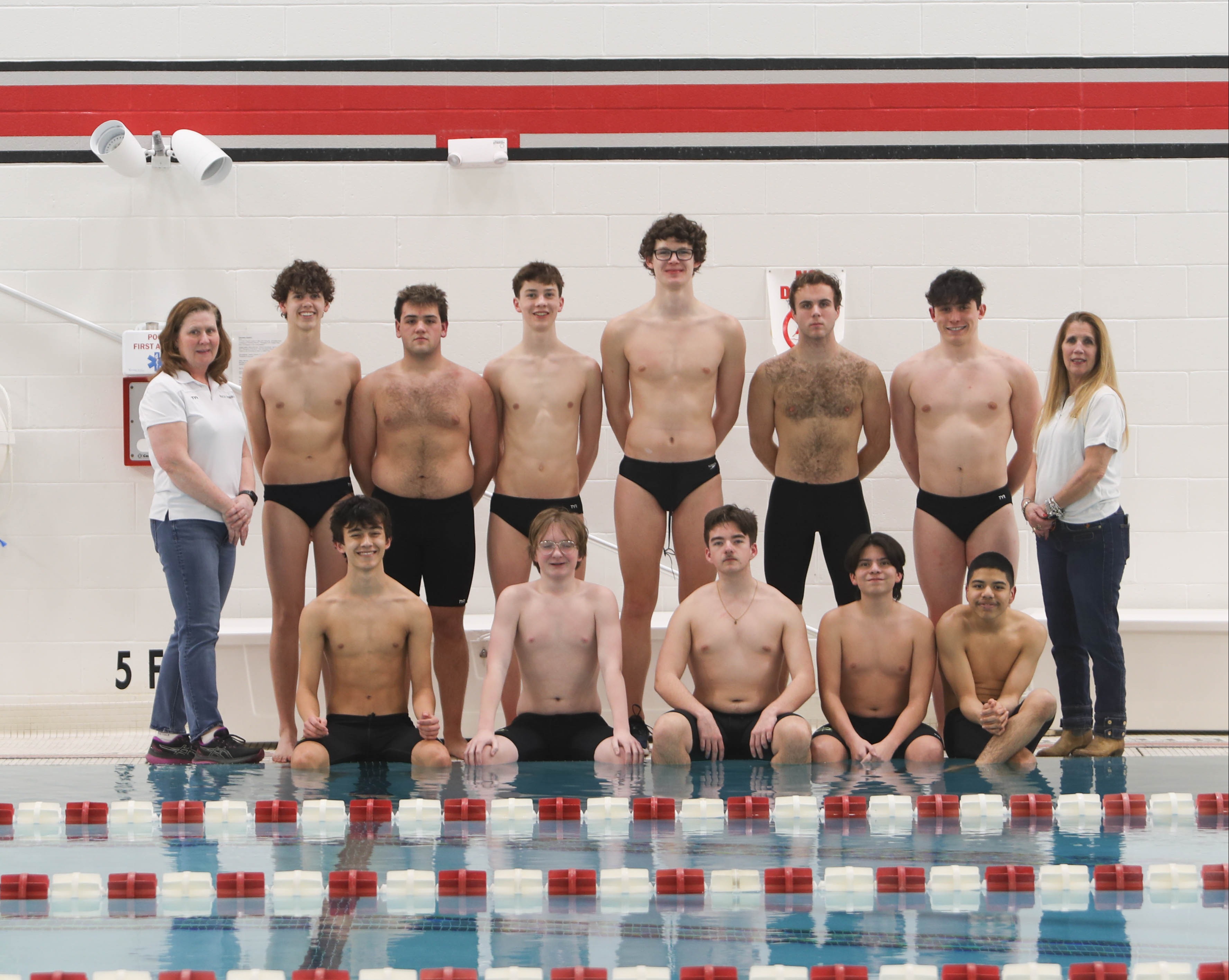 A group of male swimmers in swim trunks, wearing goggles, stand and crouch by a pool. Two women stand at the ends.