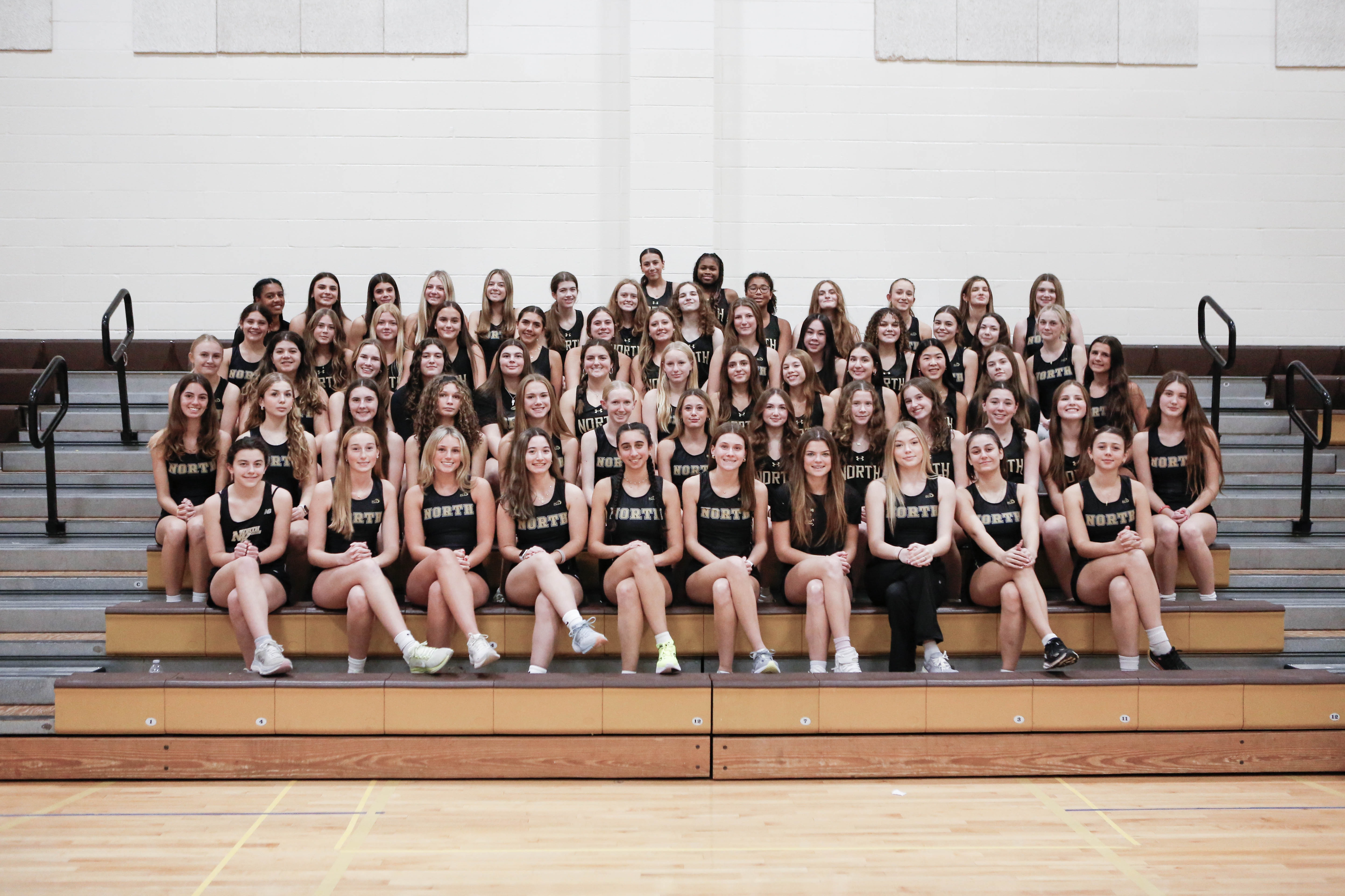 A group of athletes in matching uniforms sits and stands on bleachers in a gym.