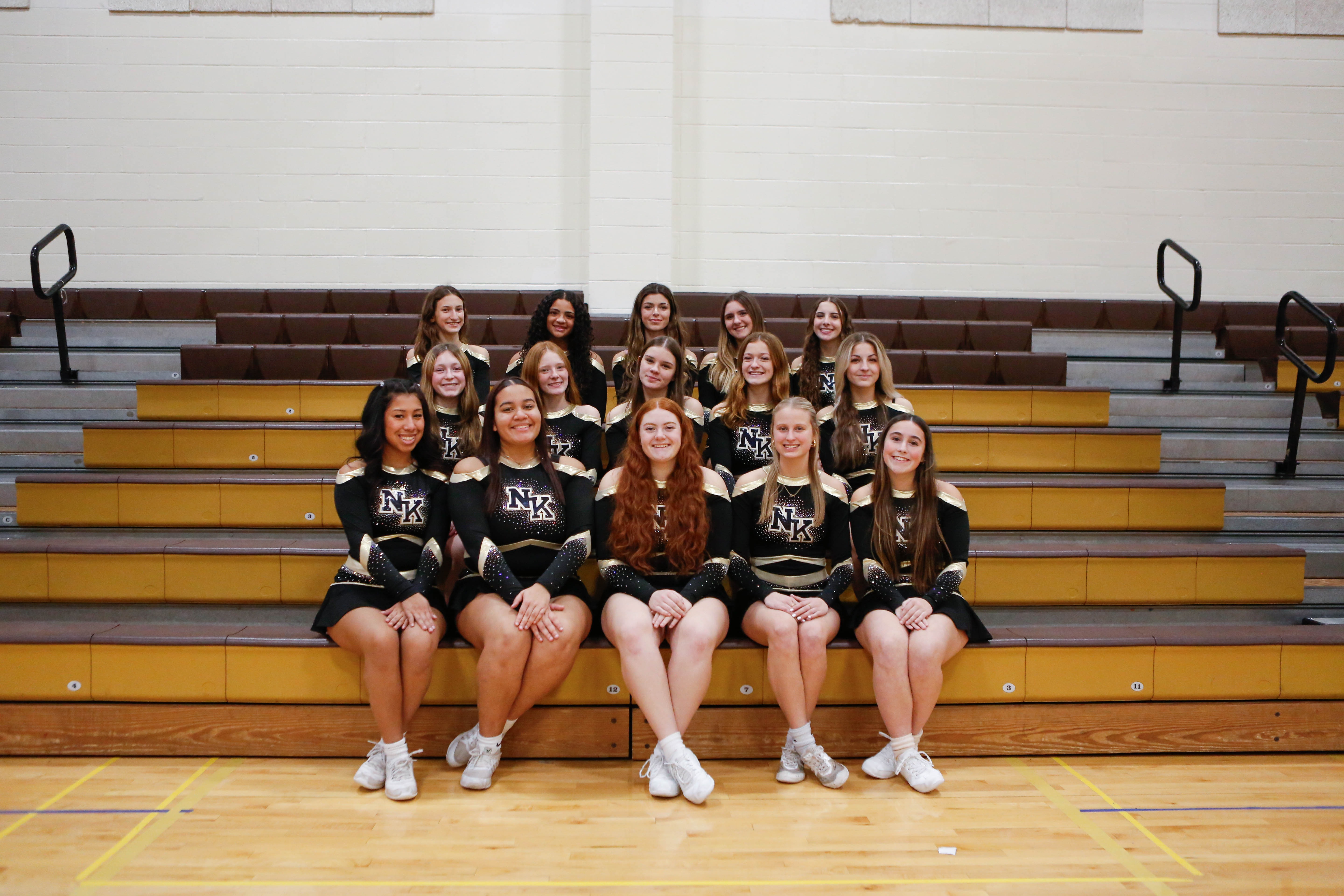 Cheerleaders in black and gold uniforms pose on a yellow bleacher in a gym with wooden floors.