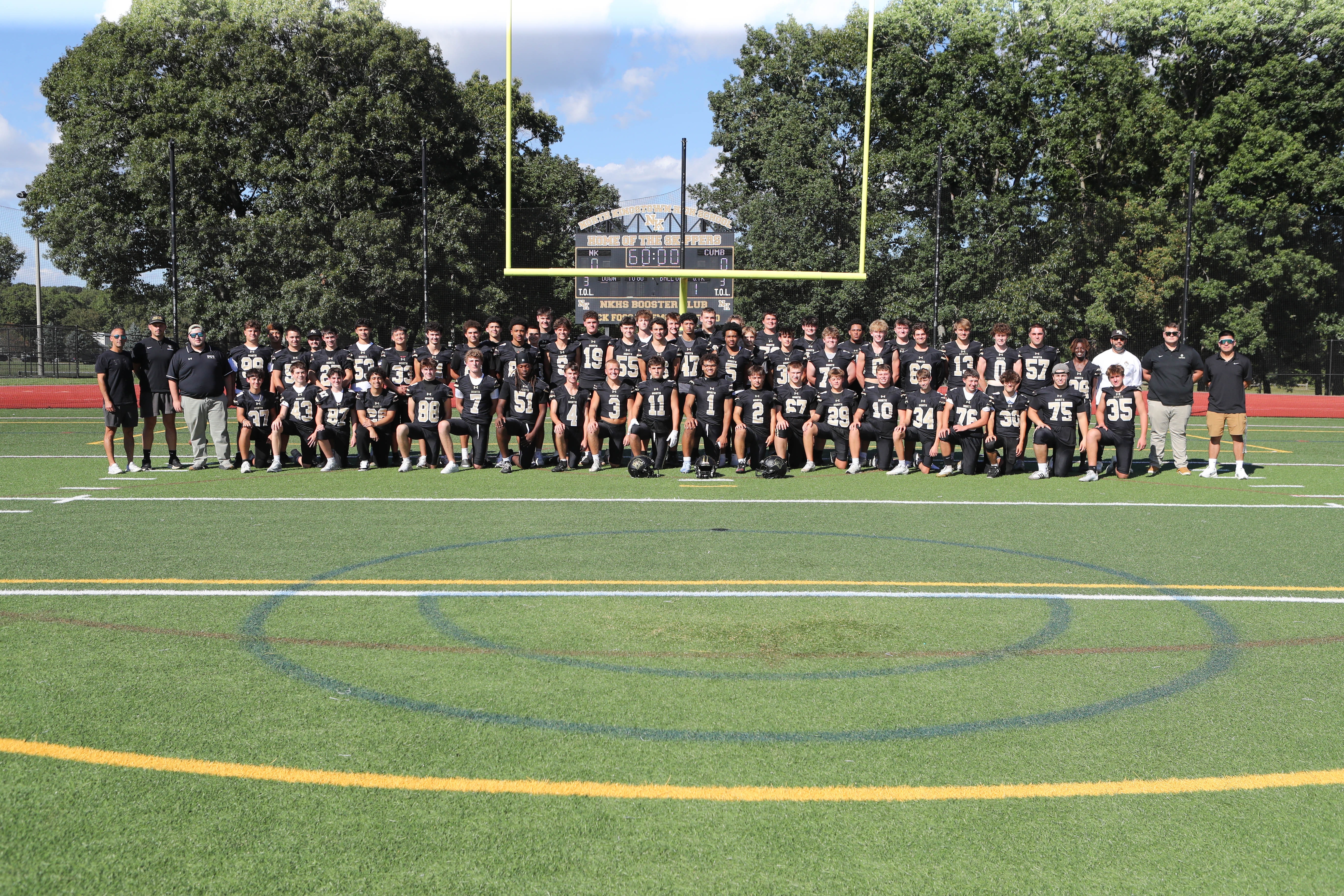 Team photo on a field with a yellow goal post. Trees and a building are in the background.