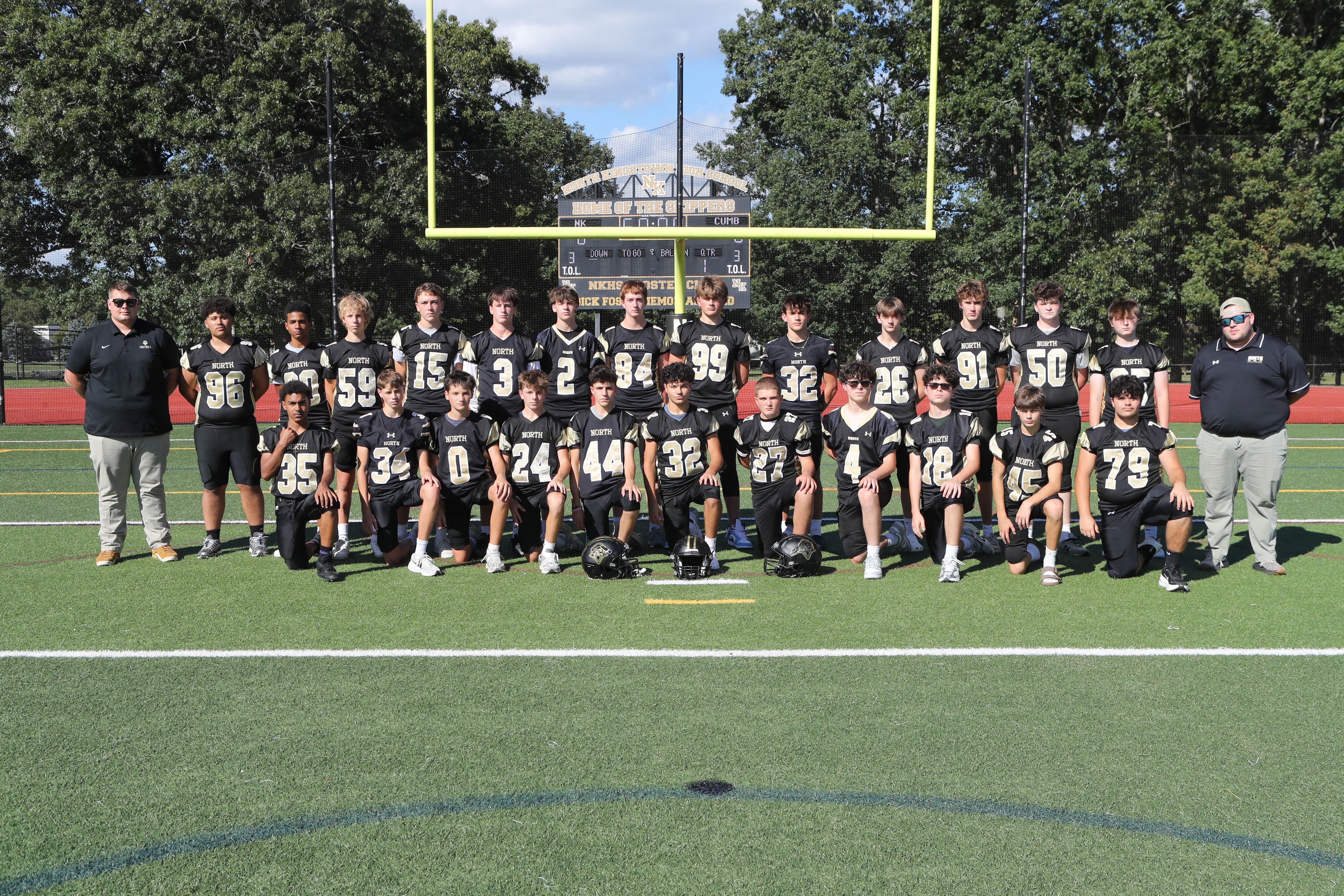 A sports team stands on a field with numbers on jerseys, two coaches on the sides, trees in the background.