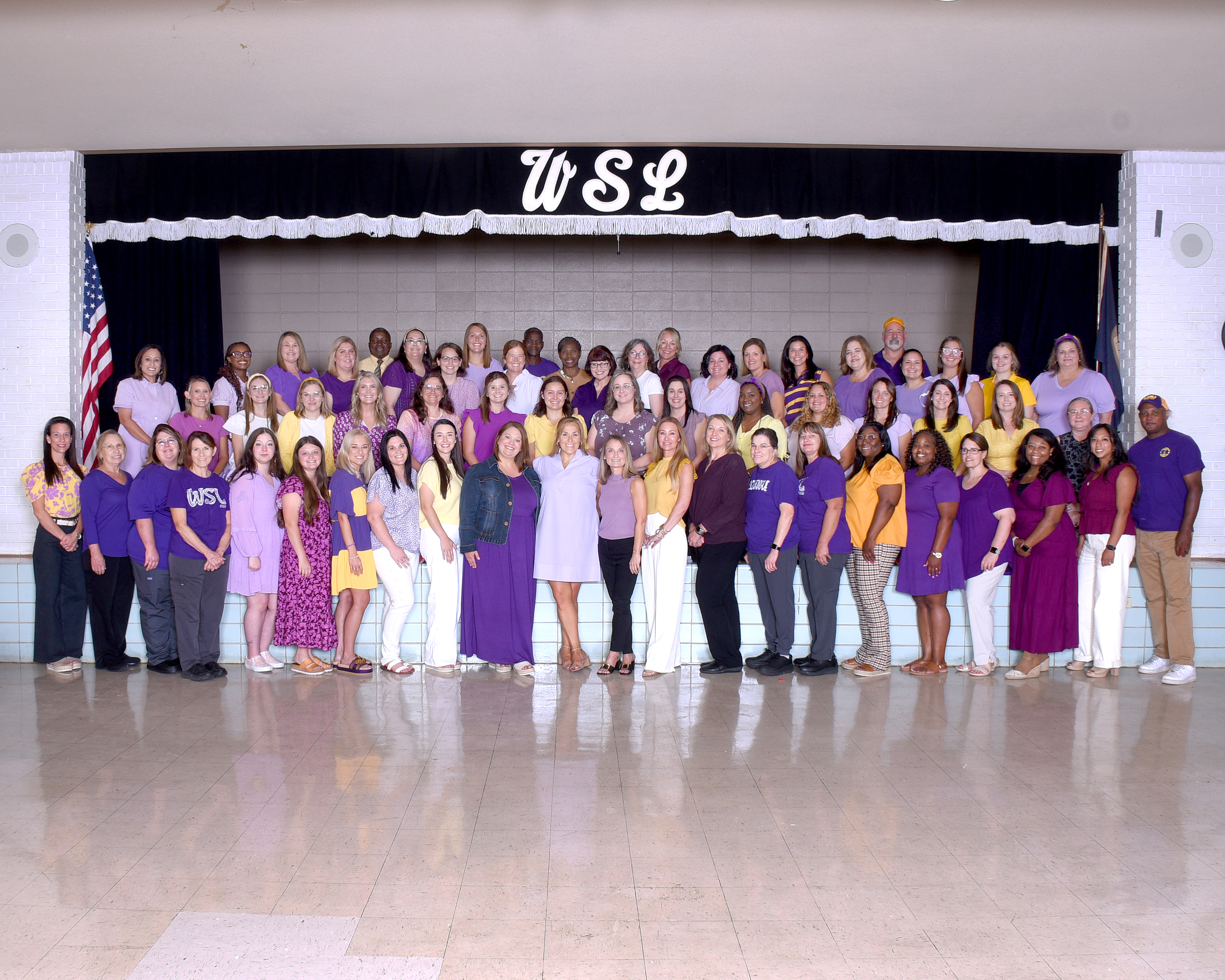 A group of adults in purple and yellow attire poses for a photo in a room with a stage.