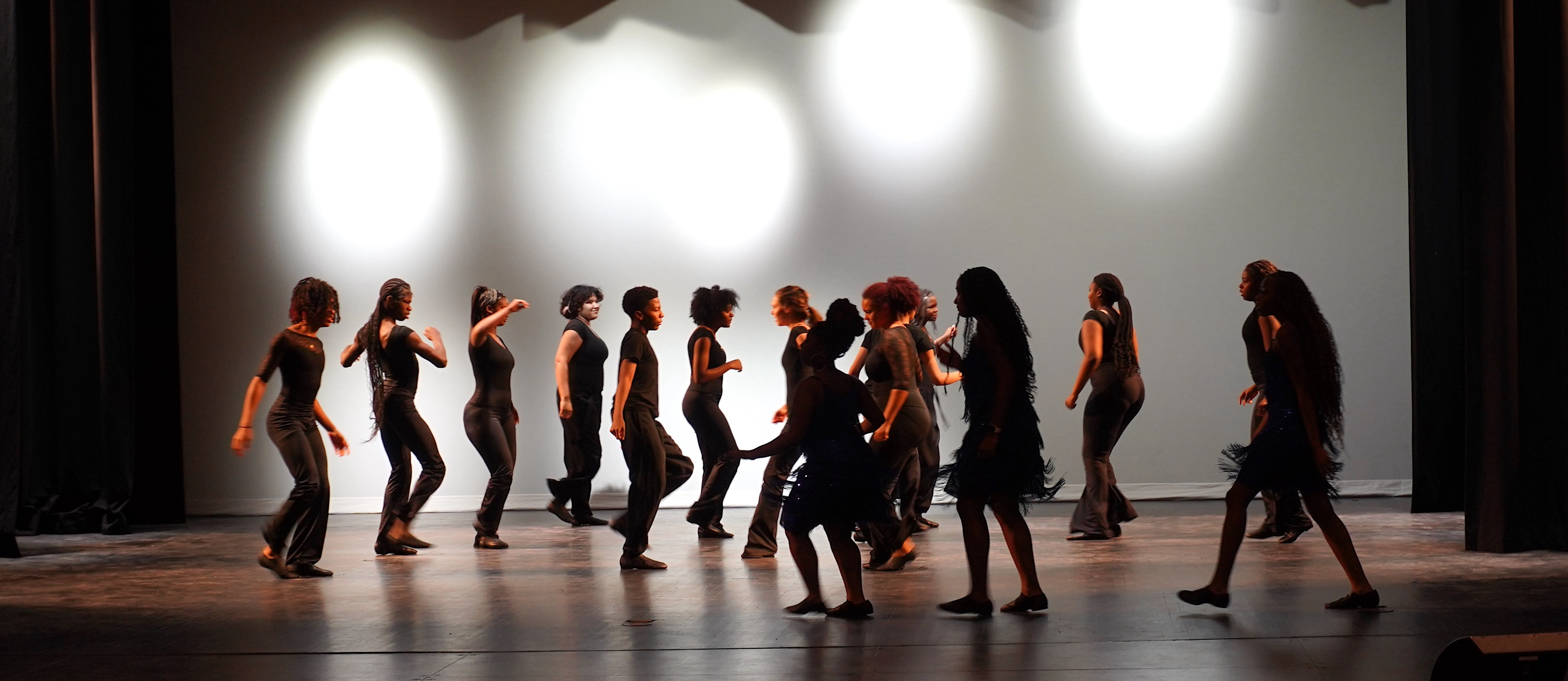 Un grupo de bailarines vestidos de negro se mueve con energía sobre un escenario tenuemente iluminado, proyectando sombras. El telón de fondo está iluminado con suaves luces blancas.