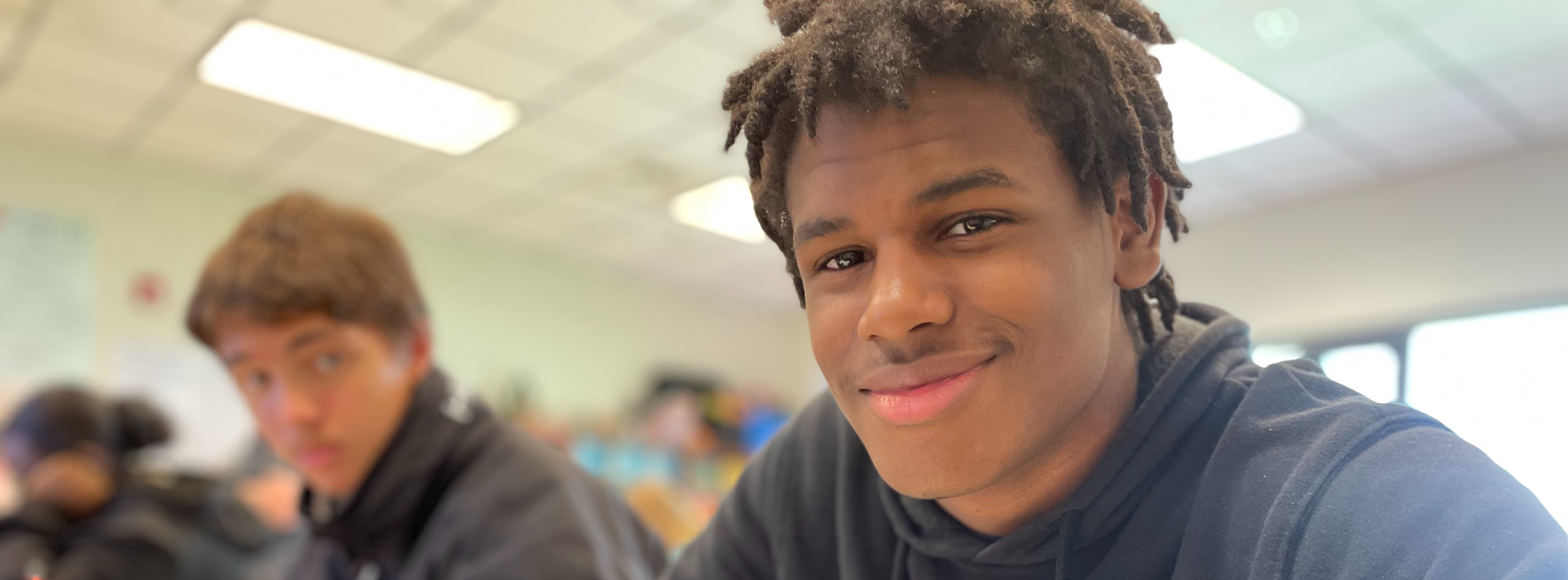 Student doing work stops to smile at the camera. Another student is sitting beside him watching.